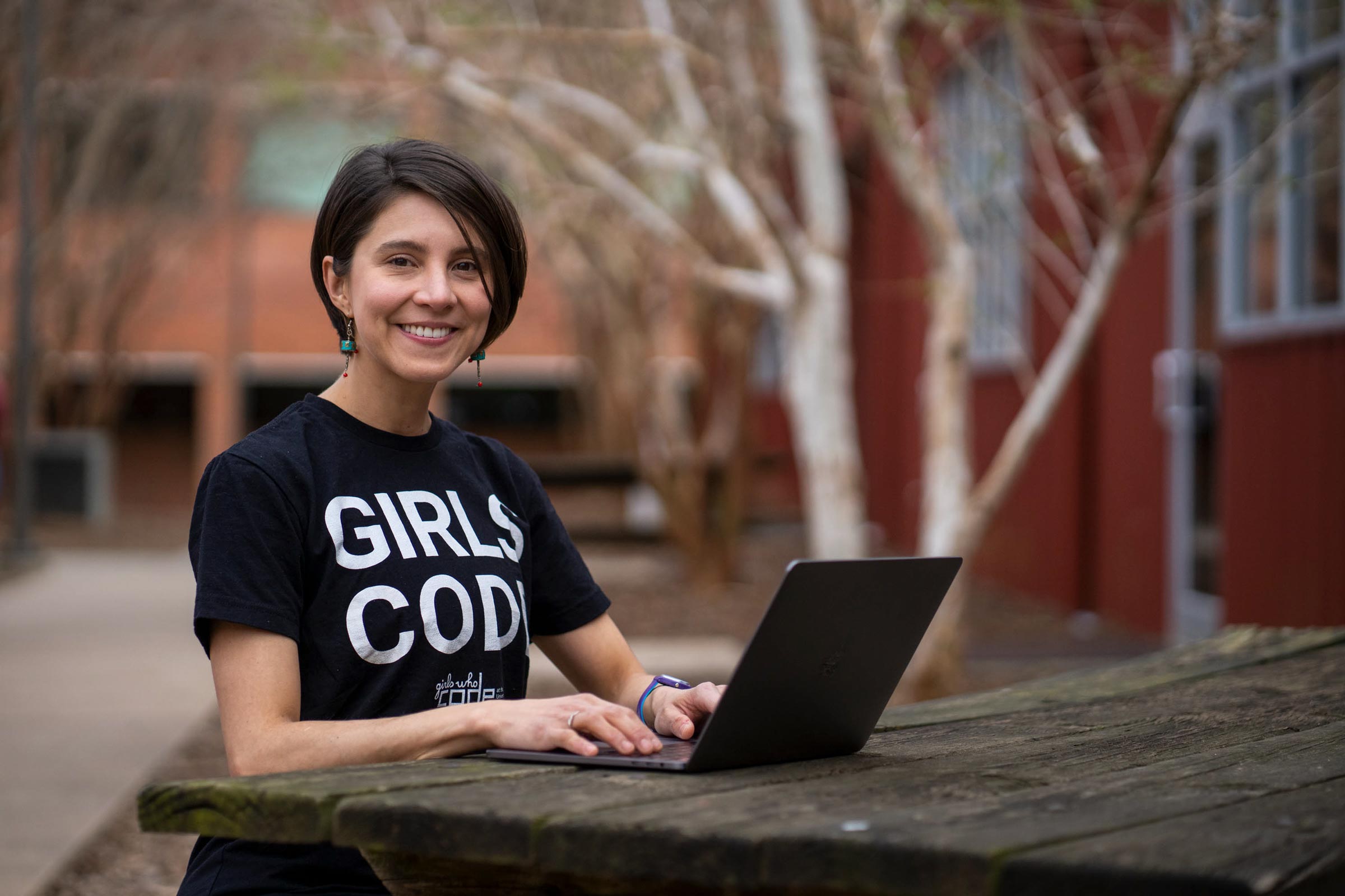 Karolina Naranjo-Velasco sits in front of a laptop on a picnic table and smiles at the camera. She is wearing a t-shirt which reads Girls Code