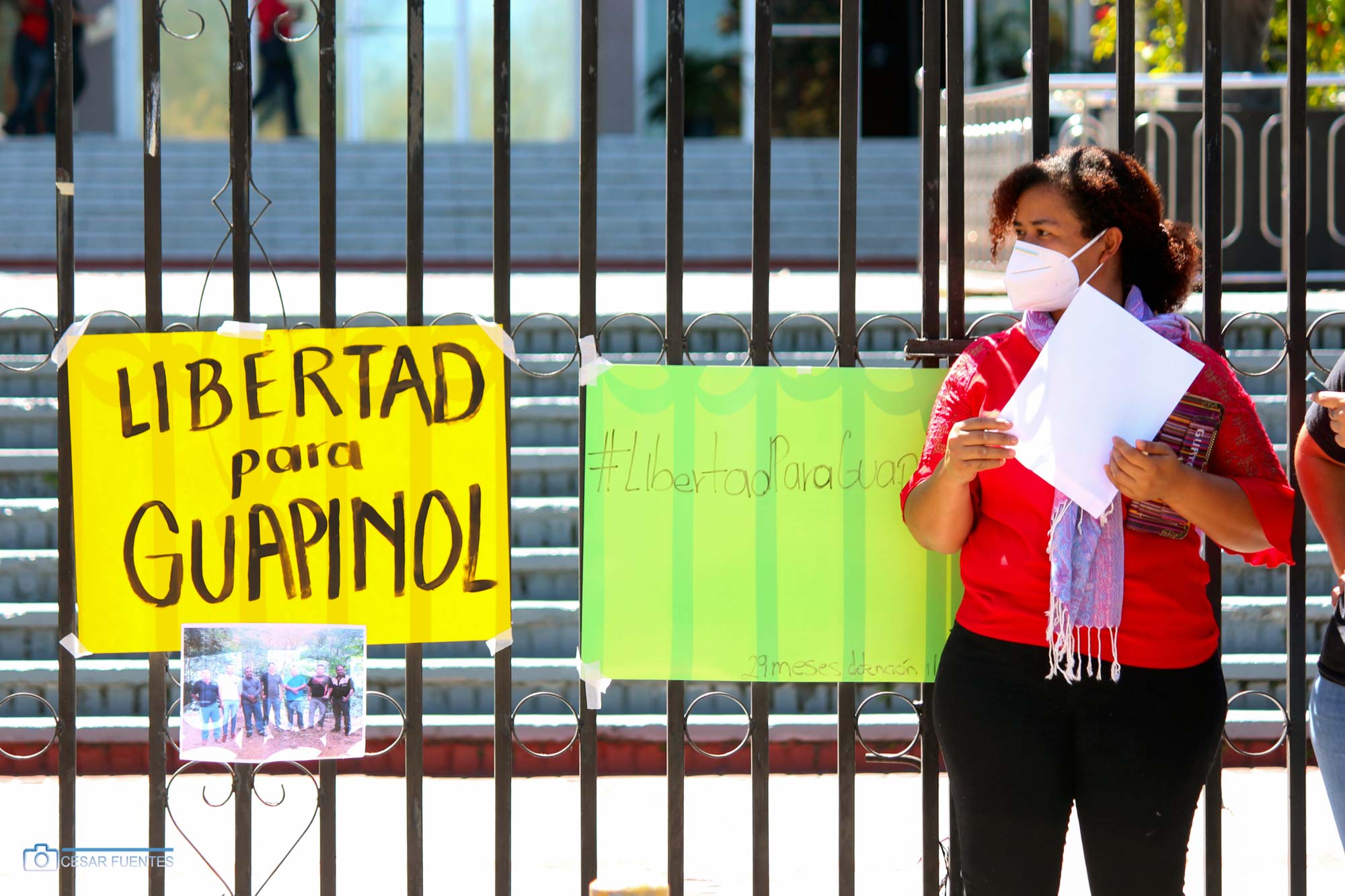 Woman standing next to a fence and sign that says Libertad para Guapinol and #LibertadPara Guapinol