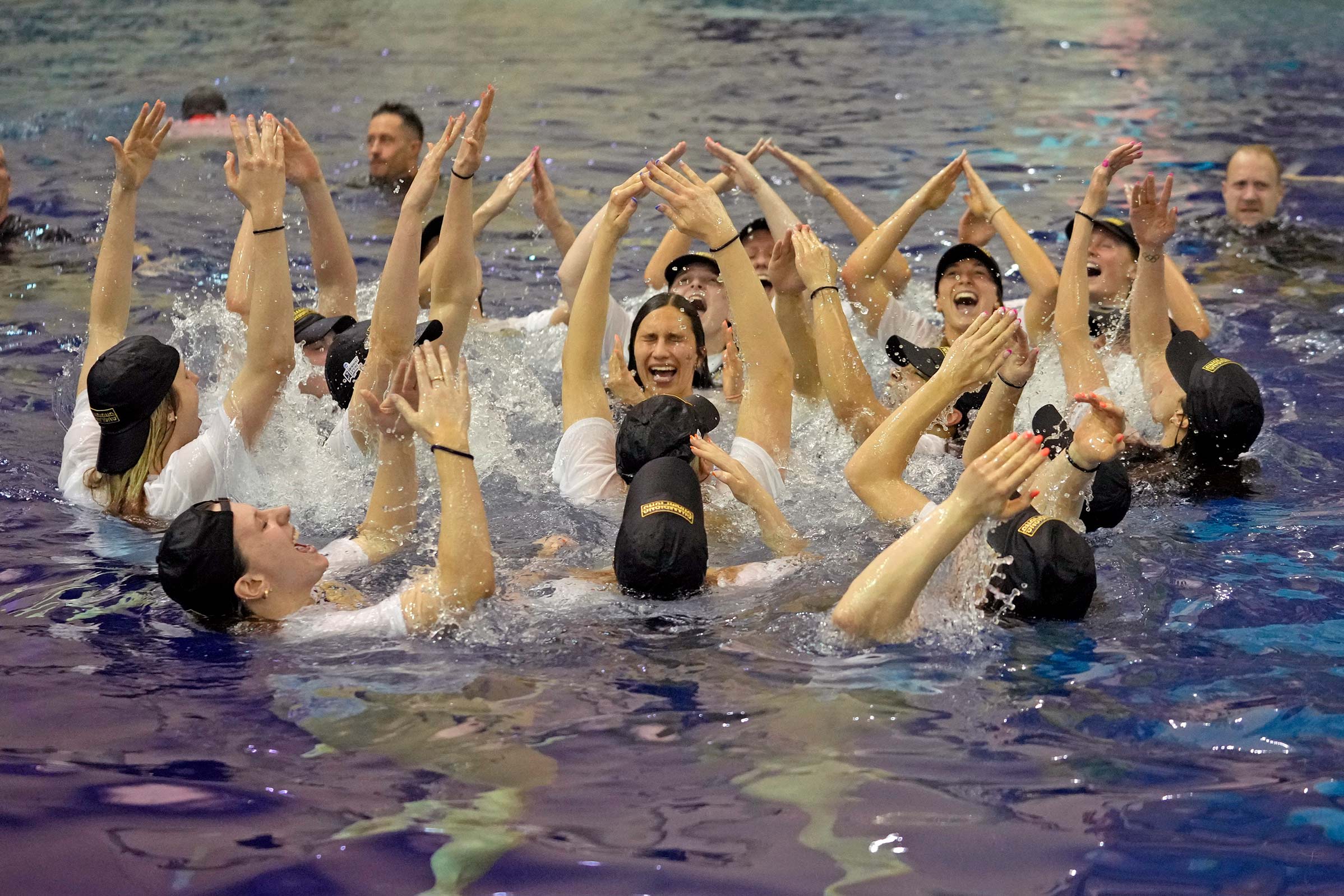 A group of University of Virginia swimmers in a pool wearing baseball caps and t-shirts smiling and raising their hands