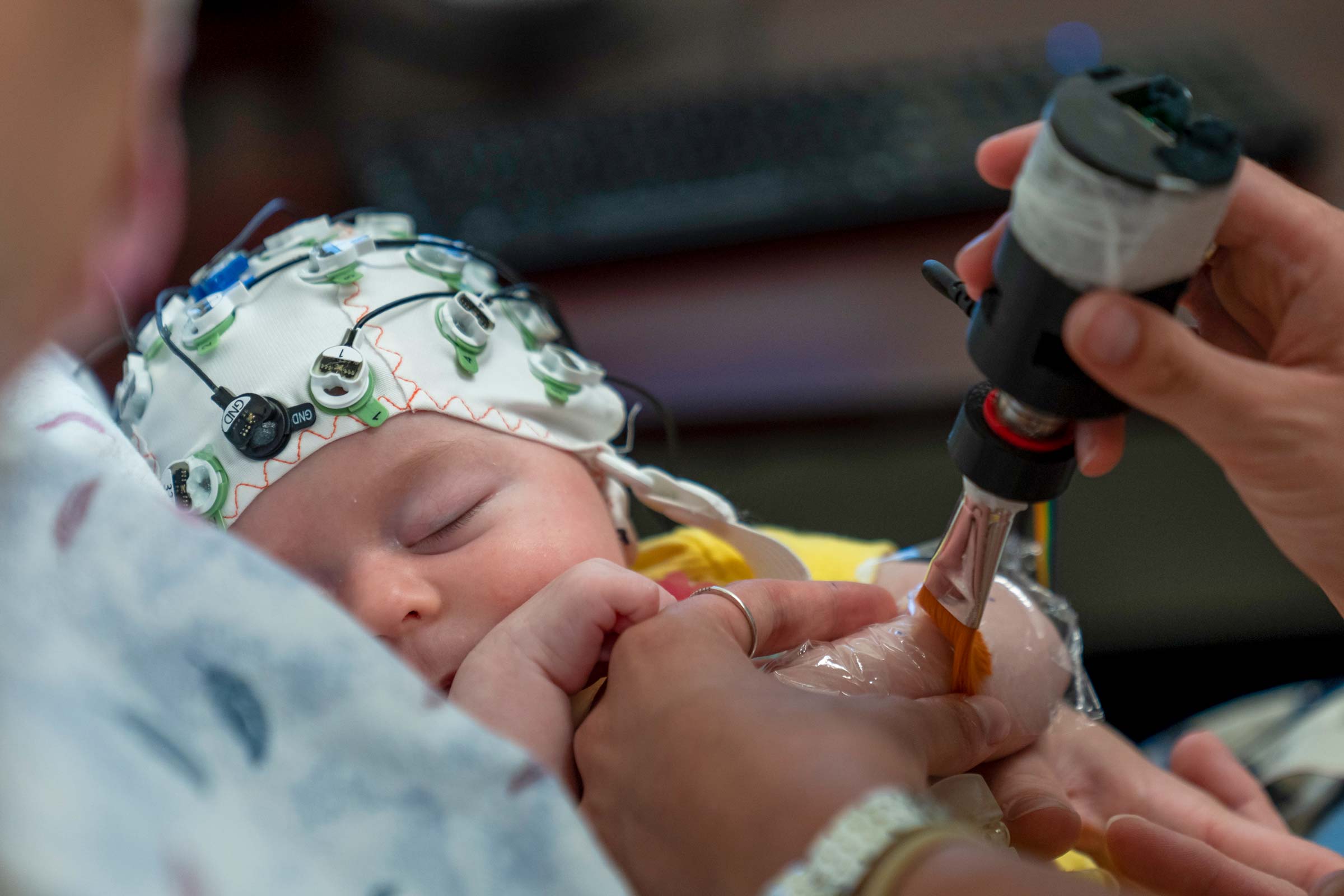 A researcher uses a paintbrush to stroke the arm of a sleeping baby, who is wearing a skullcap with lights and wires connected to it