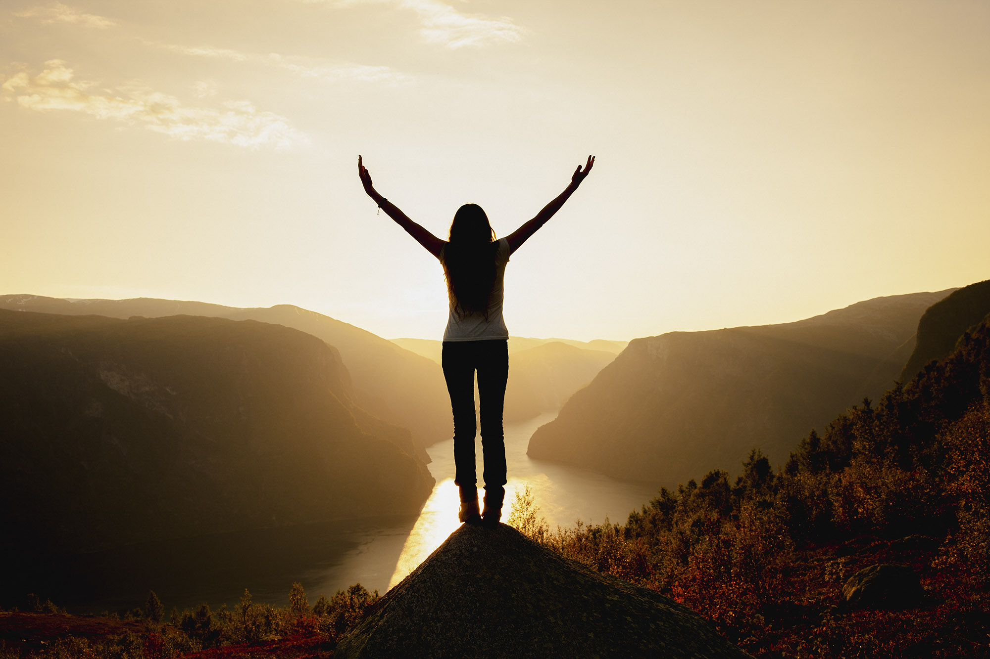 Woman standing on the edge of a mountain with arms raised over looking a river