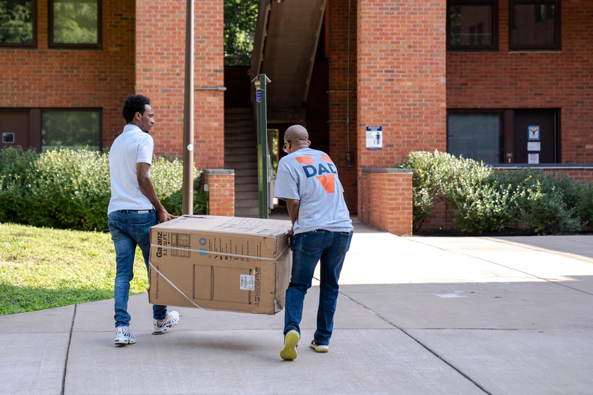 A man in a Virginia Dad t-shirt helps his son move a large box into a dorm