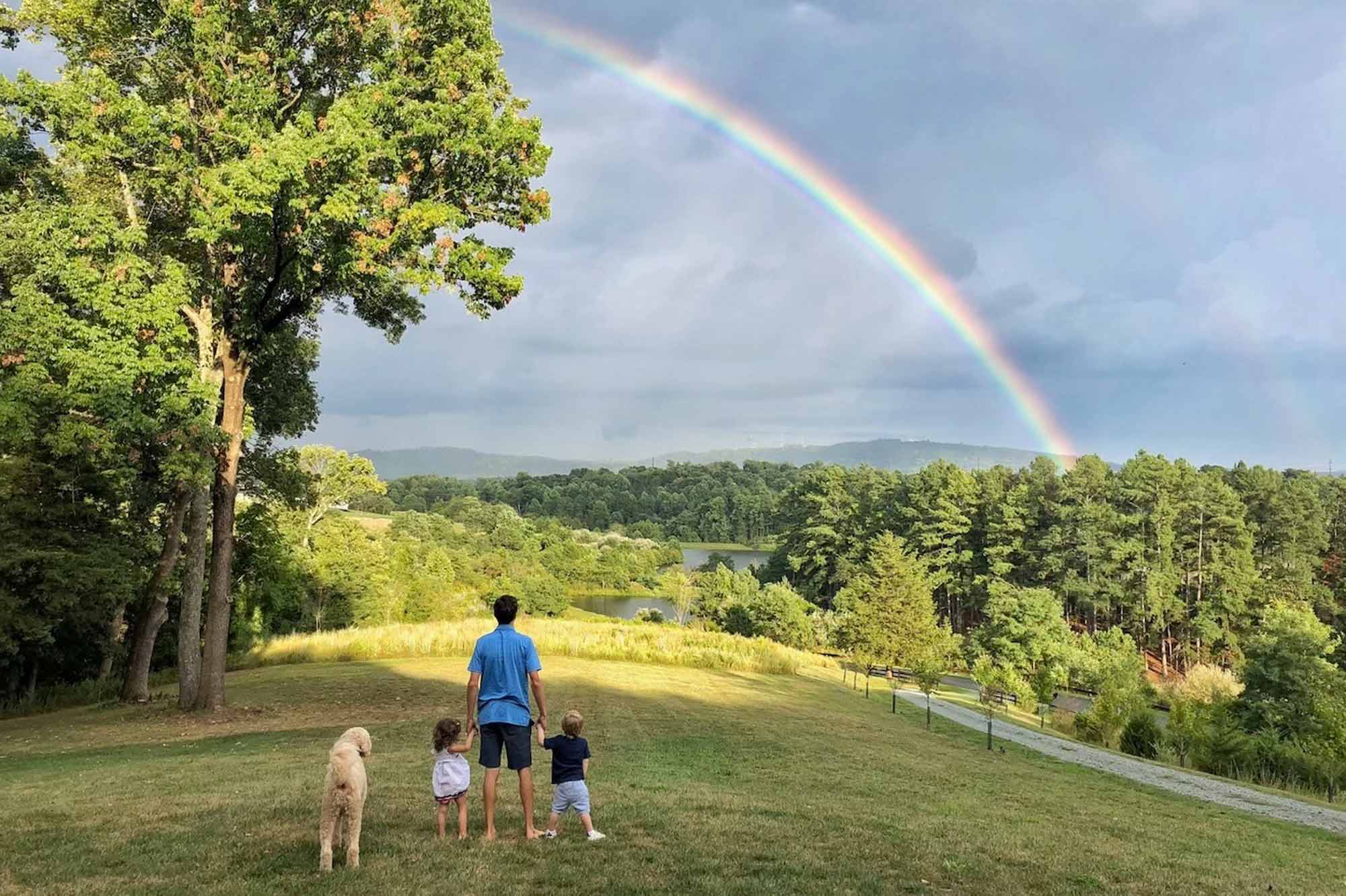 Father, two kids, and the family dog stand on a hill looking up at a rainbow
