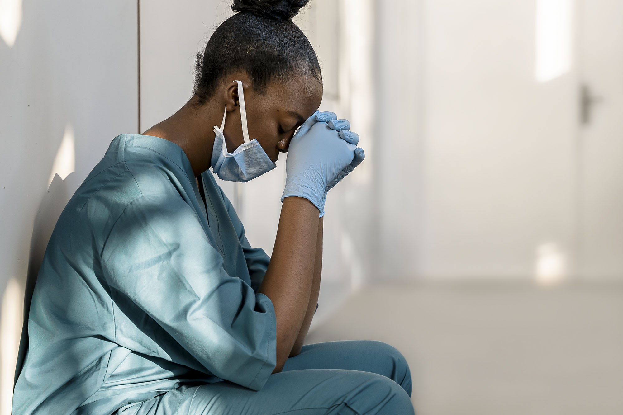 Nurse with elbows on her knees and head on her hands wearing a mask in a hospital hallway