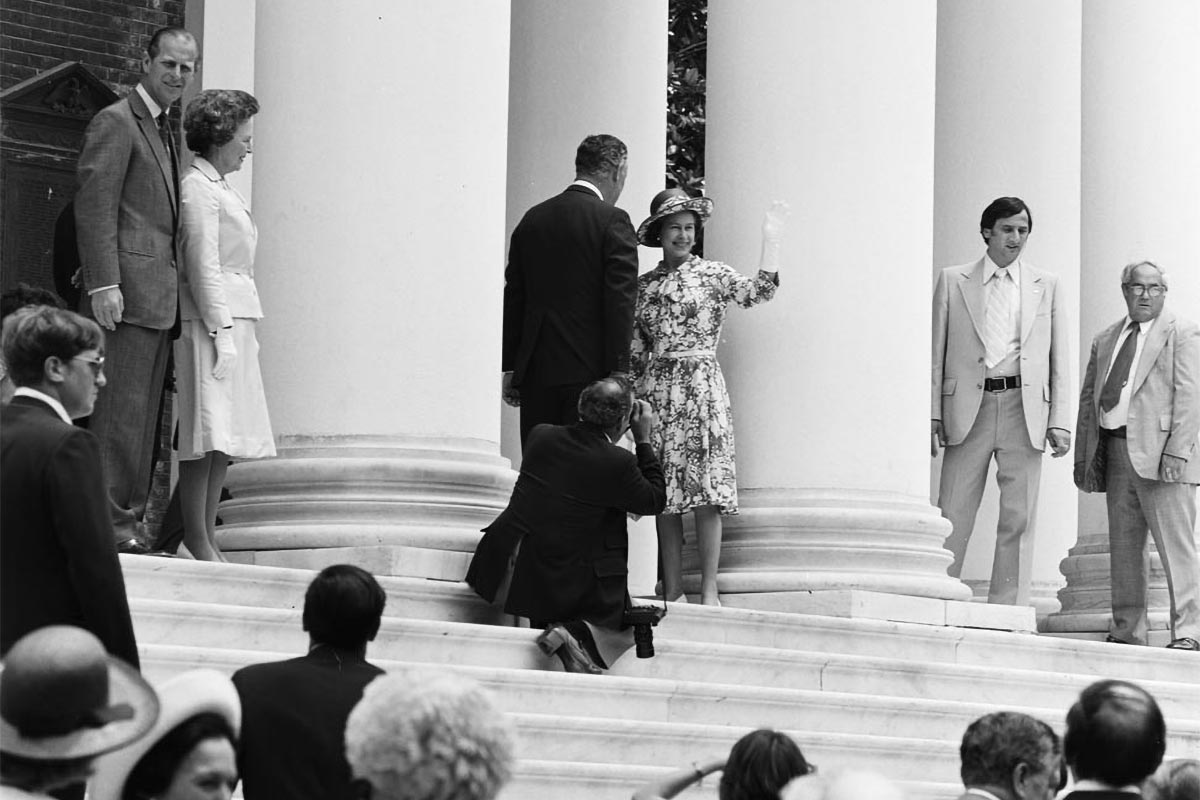 Queen Elizabeth, in a floral dress, gloves and a hat, stands at the top of the Rotunda steps and waves at a gathered crowd