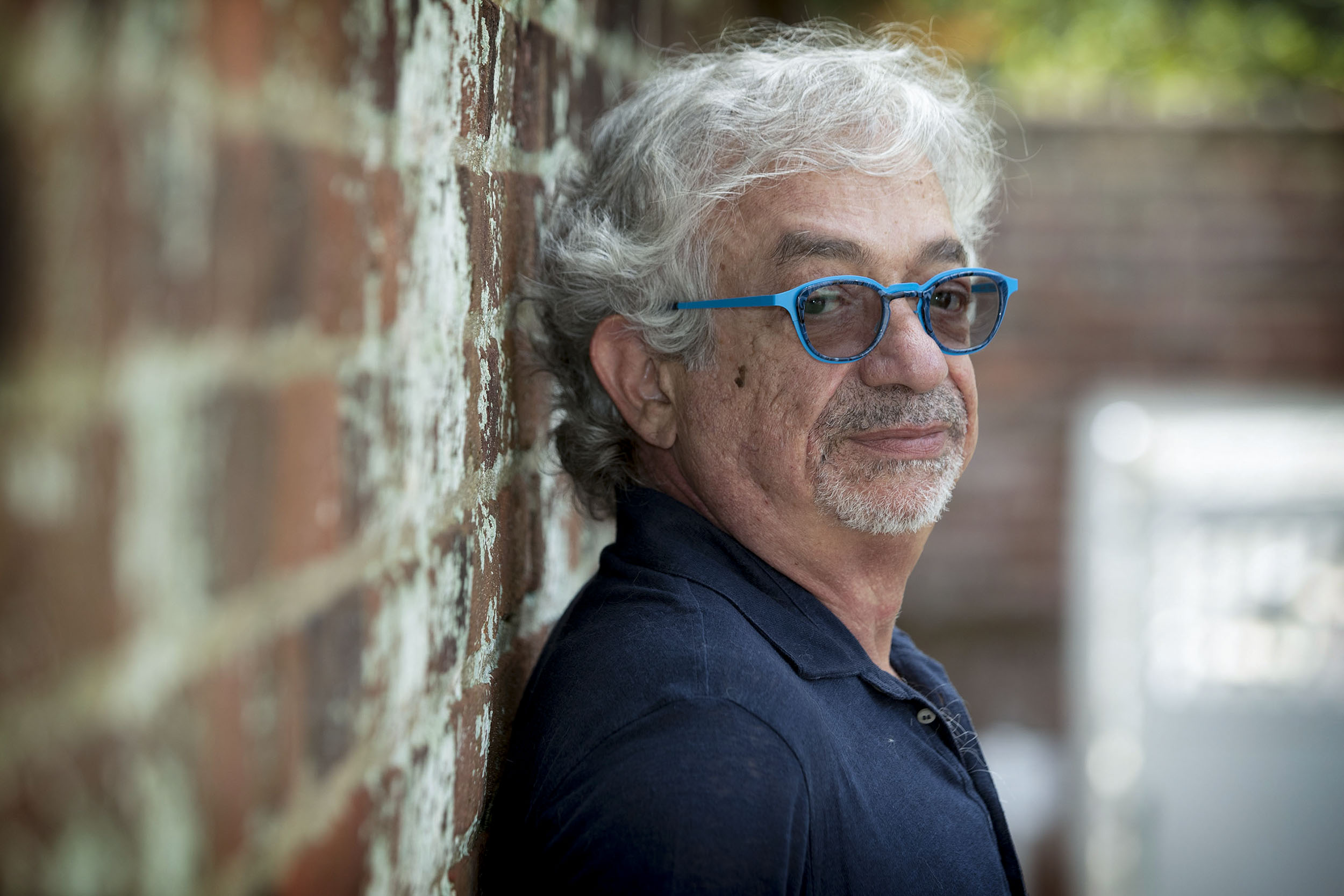 A man with blue glasses and a goatee leans against a brick wall