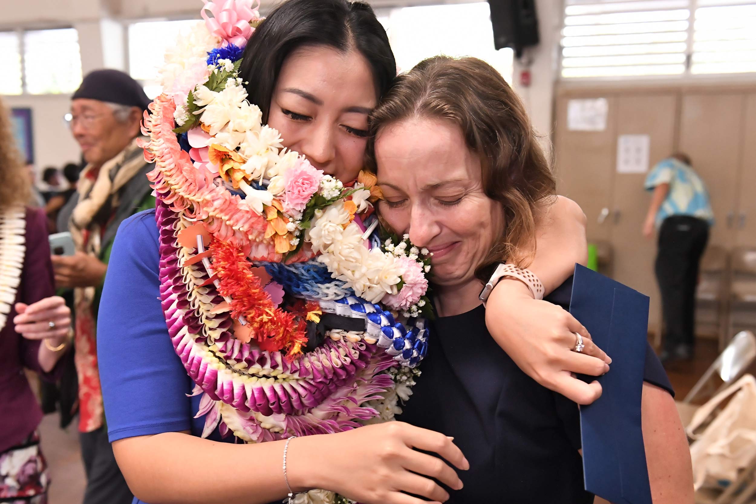 Esther Kwon coverd in ceremonial leis hugging a colleague in her elementary school