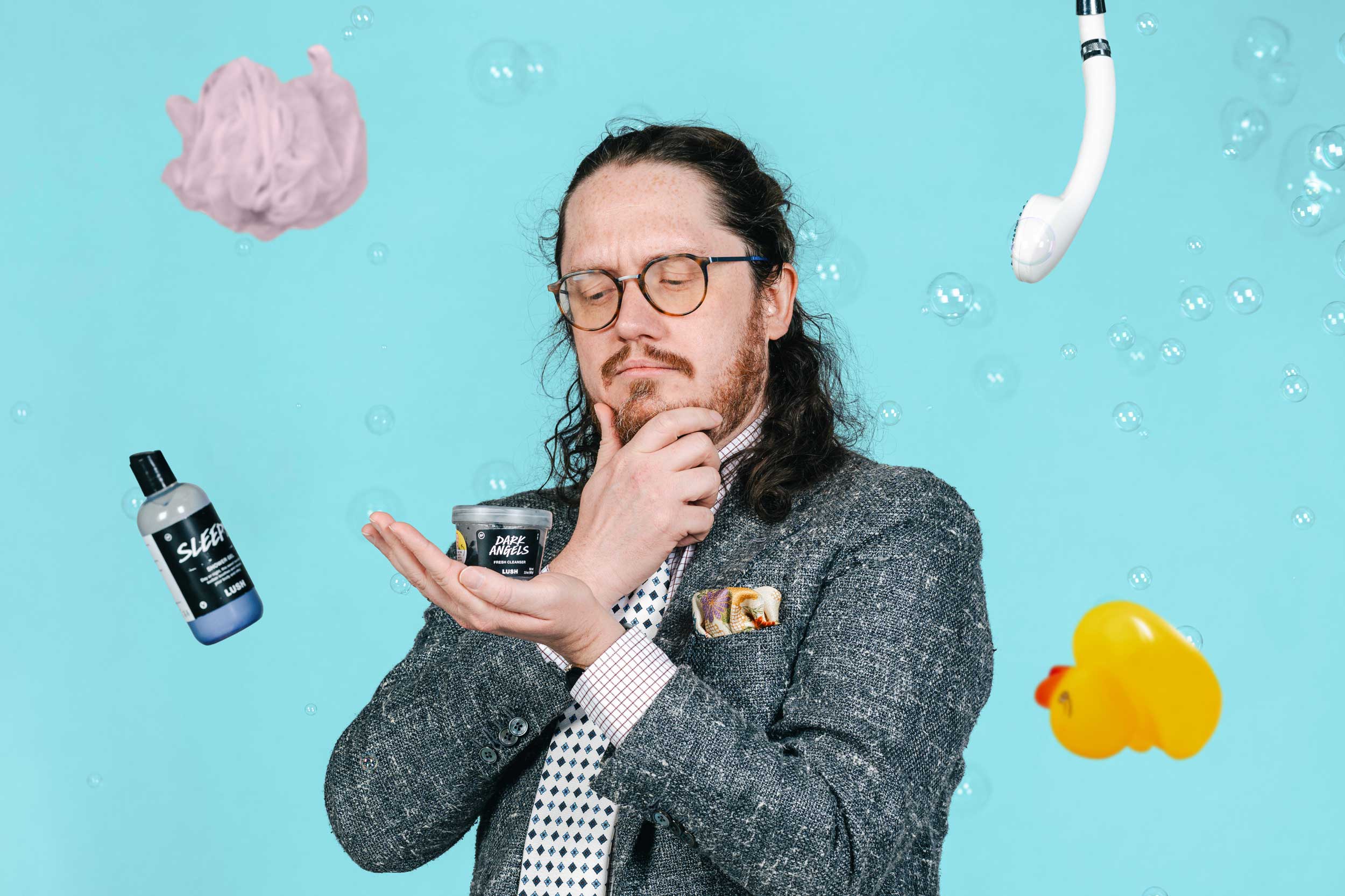 Zachary Irving looks at a "Lush" product with a pensive expression while other shower items hang in the air around him