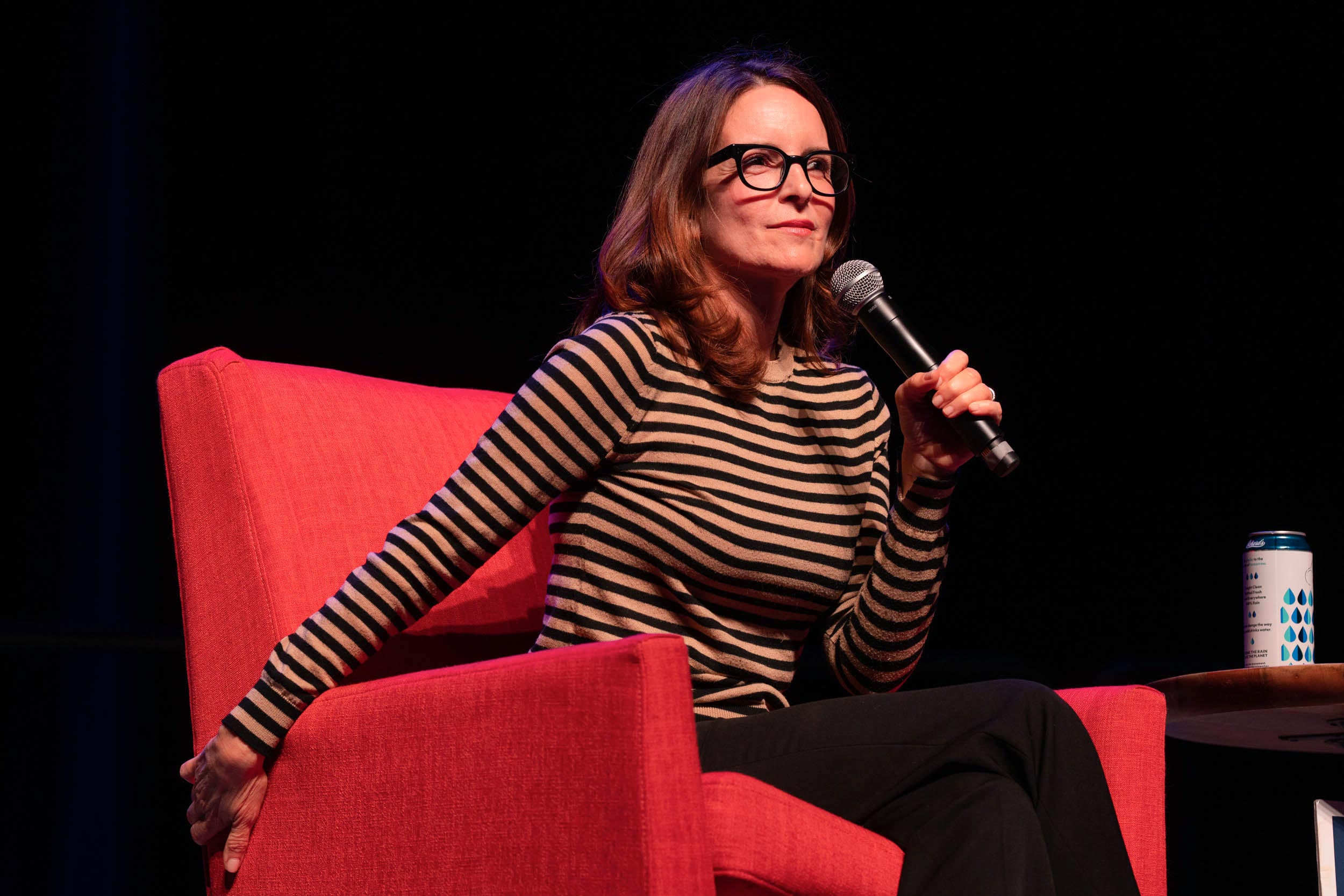 Candid photo of Tina Fey on stage sitting in a red chair holding a microphone