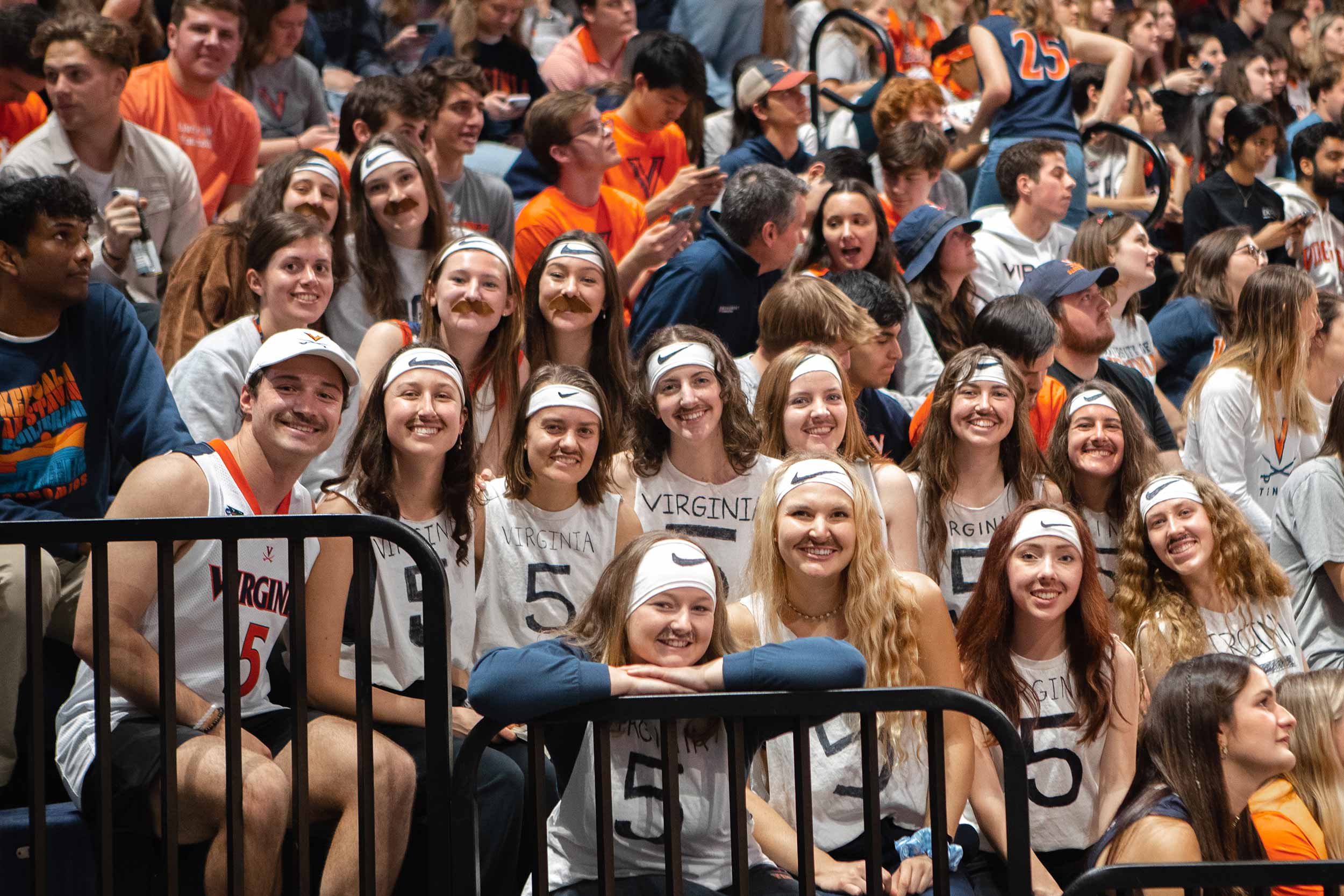 A group of students wearing fake mustaches and white sleeveless teeshirts with the number 5 written on it at a UVA Men's Basketball home game