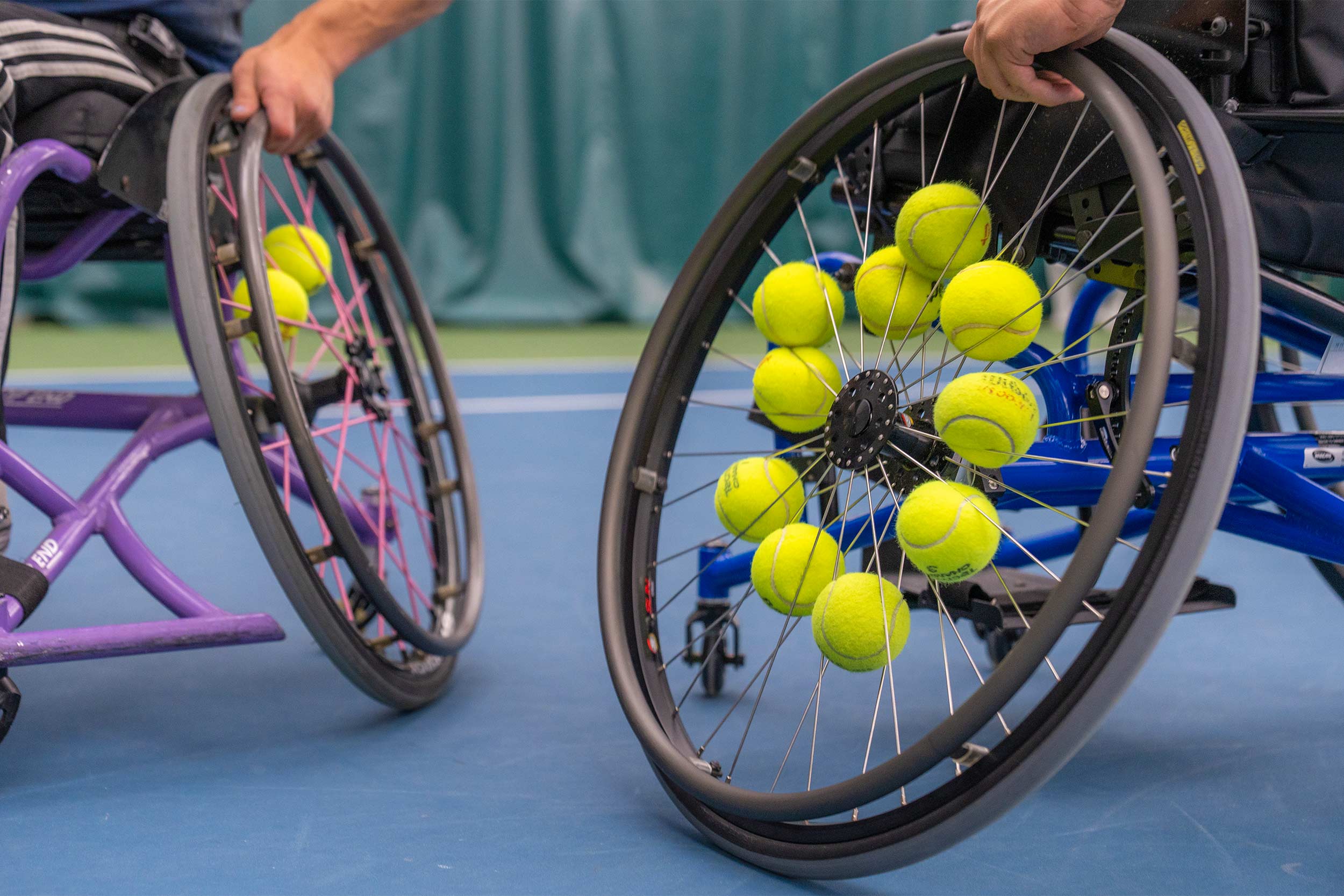 Two wheelchairs, with tennis balls in their spokes, rest on a blue tennis court. 