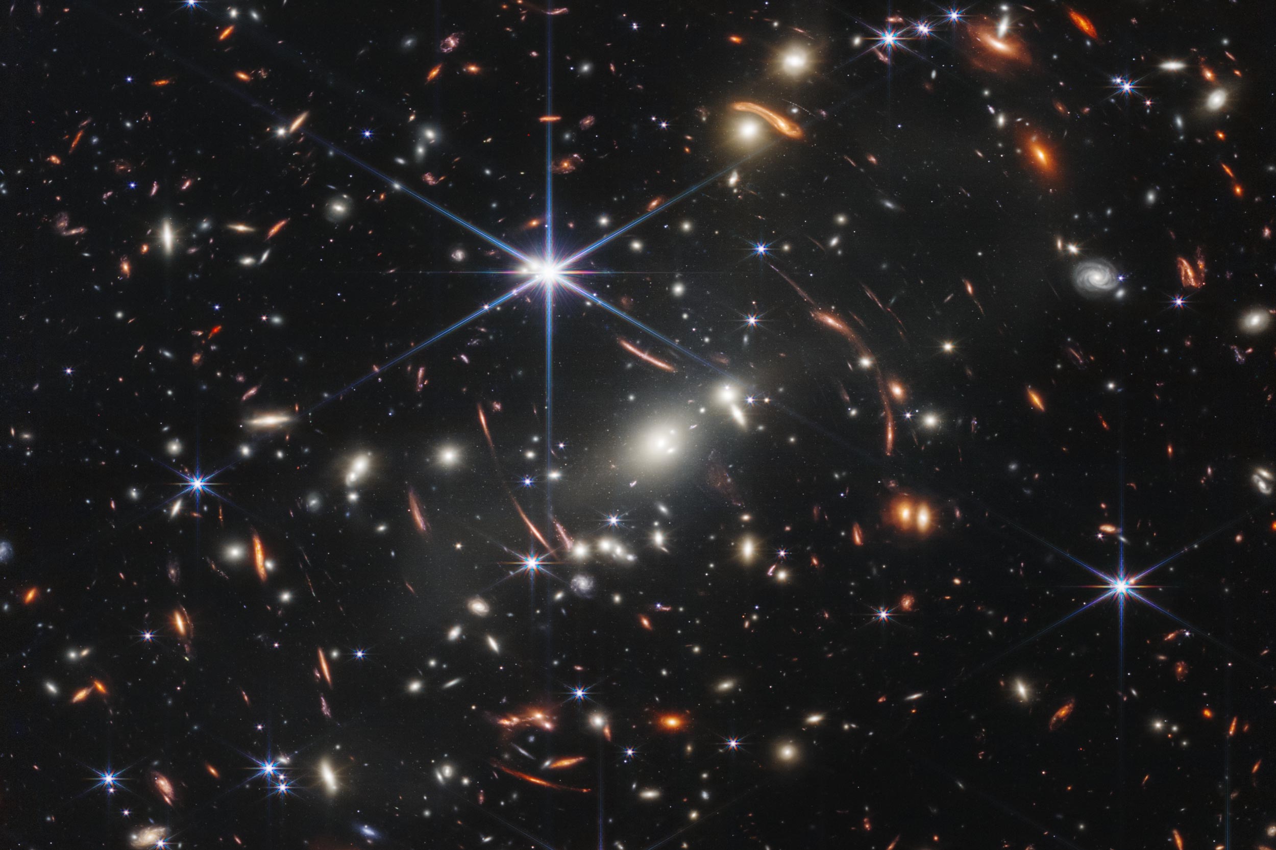Hundreds of galaxies, including dozens of curved red-orange lines, bending around two bright, white oval galaxies in the center