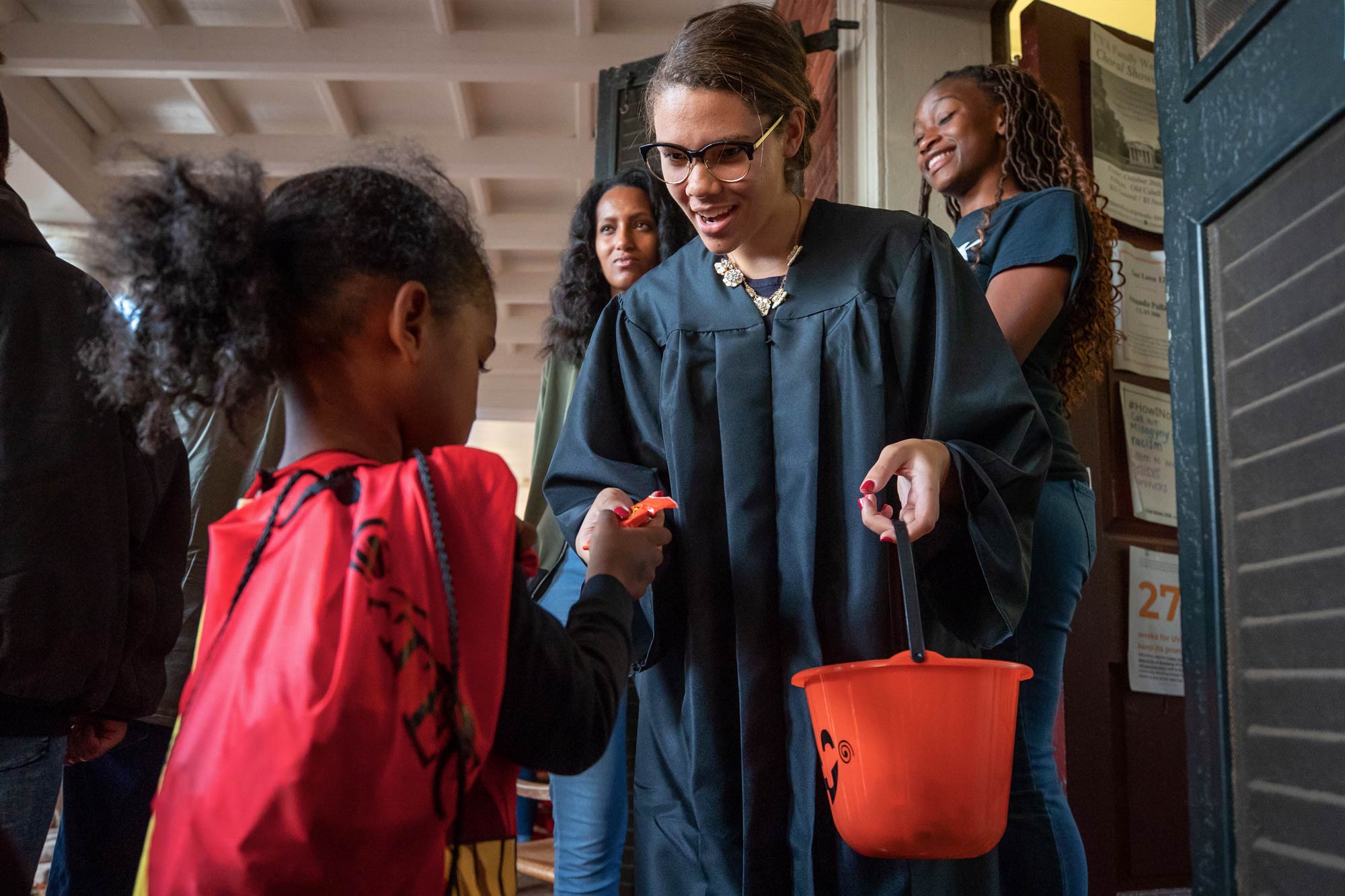 A college student in a judge's robes and glasses gives candy to a small child in a red cape