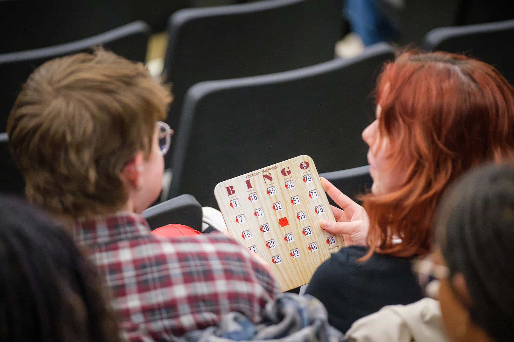 Over the shoulders of two students holding a reusable wooden bingo card with slides to go over each number