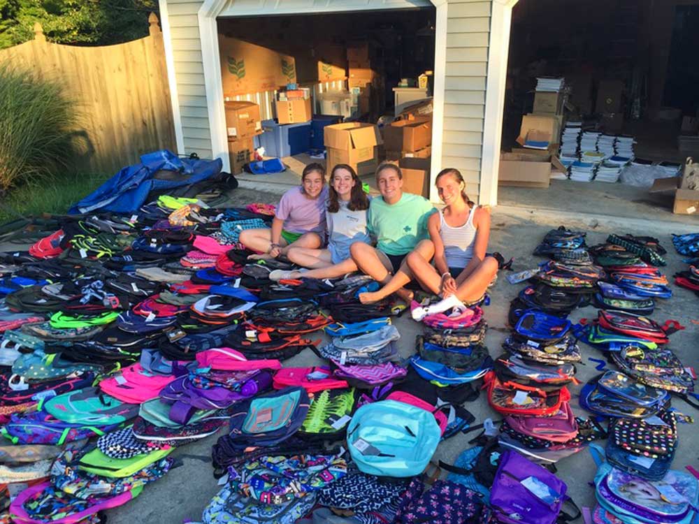 Lucia Hoerr Jayne Frazier, Emily Trebour and Molly Morris sitting in a driveway surrounded by backpacks with a garage full of school supplies