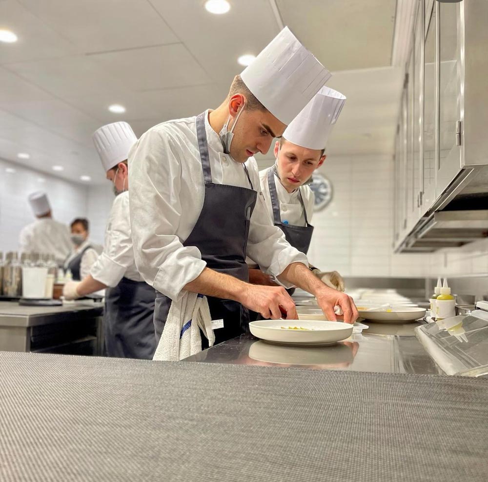 Portrait of Verheggen learning a technique from another chef in the kitchen.