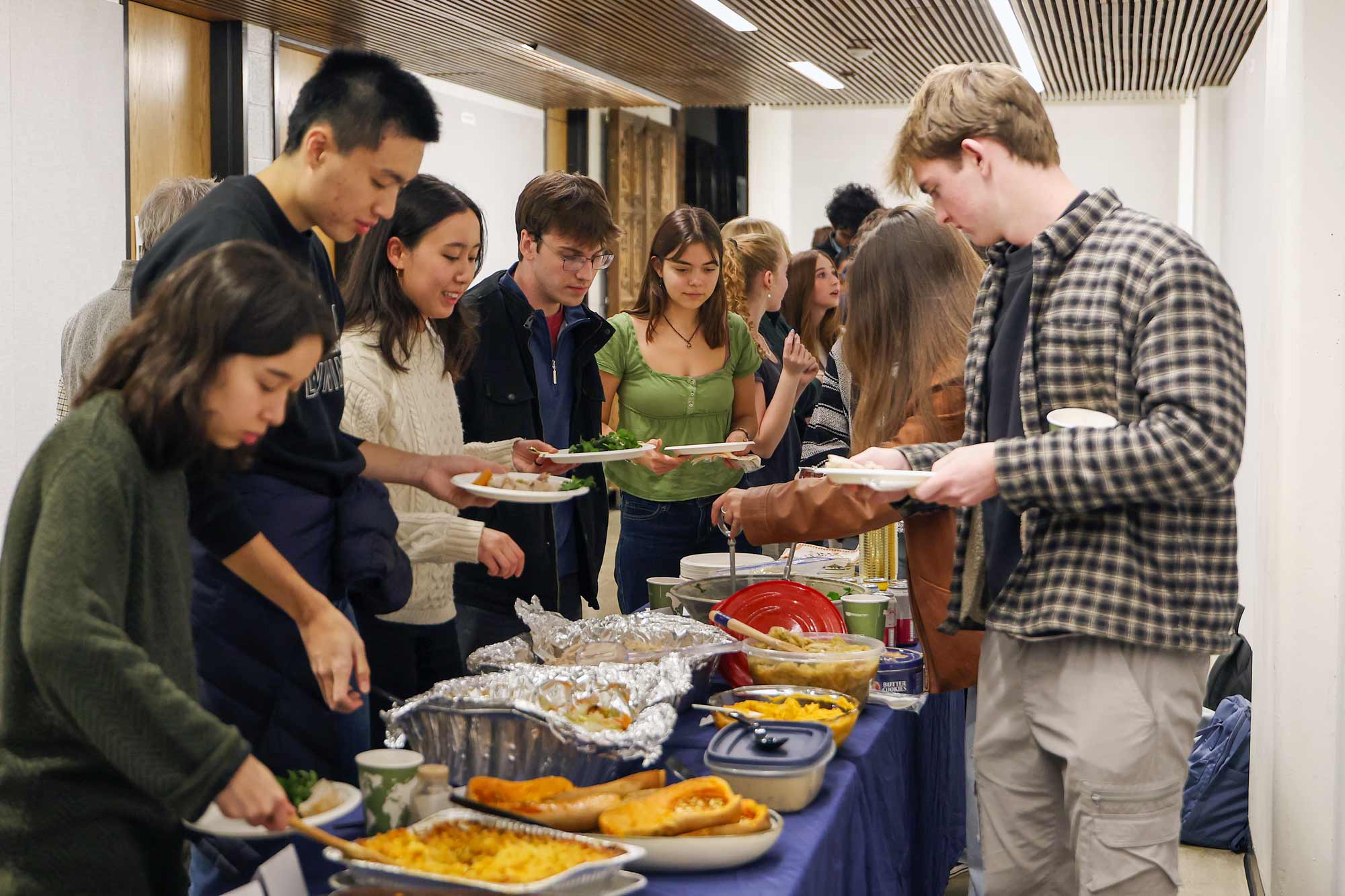 Architecture students serving themselves food from a long buffet table at a communal meal, with various dishes laid out and guests holding plates as they move through the line.