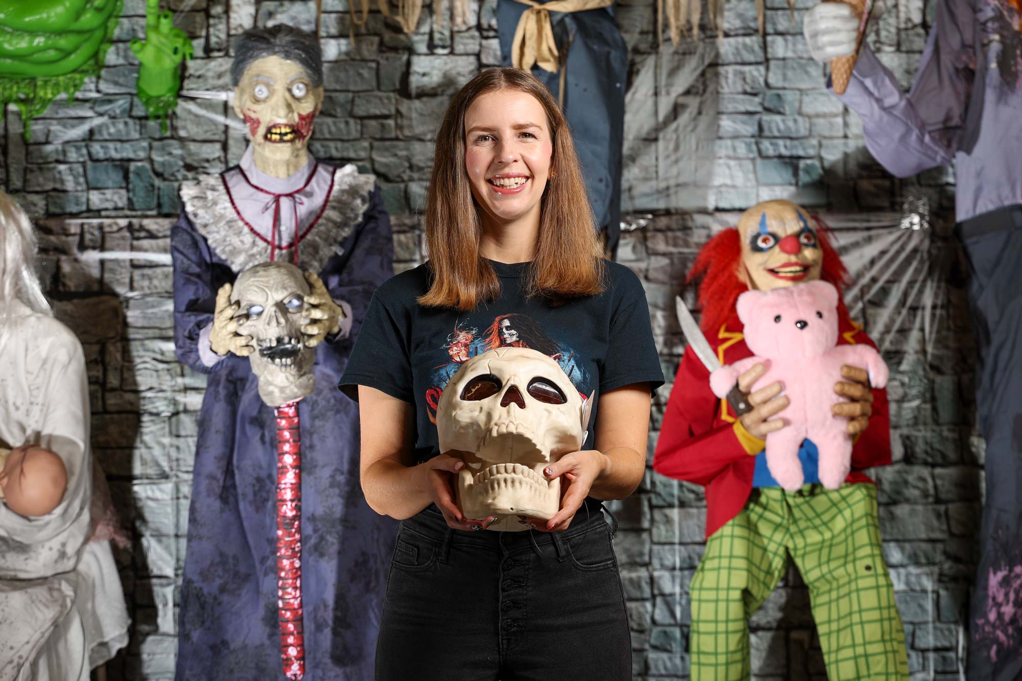 Natasha Bailey holding a novelty plastic skull in front of several halloween decorations including a hag holding a skull and spine, and a scary clown holding a stuffed teddy bear and a large knife.