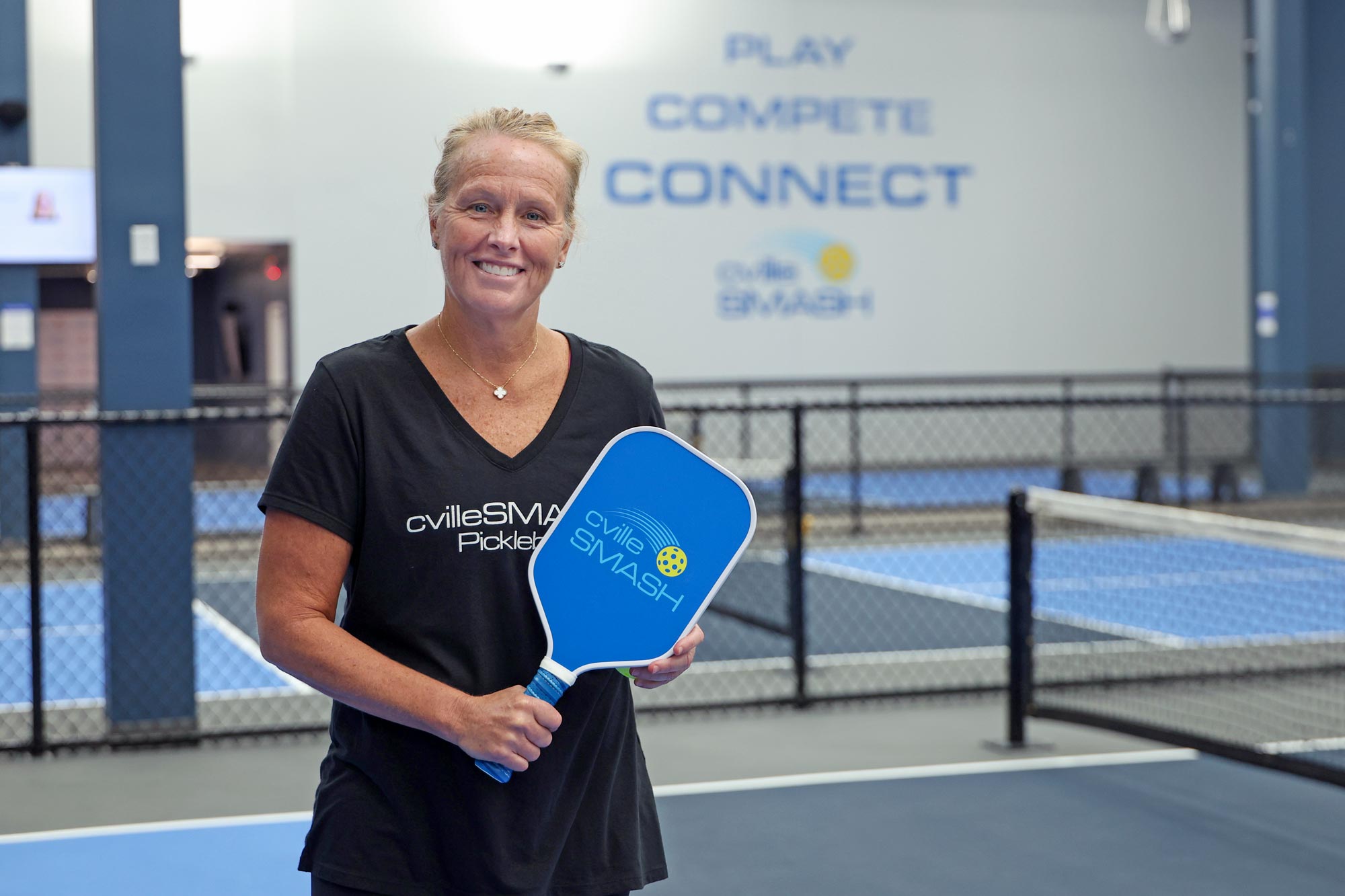 Colleen Shearer posing in the new Cville Smash pickleball facility. The far wall says “Play. Compete. Connect” and she is holding a paddle with the Cville Smash logo on it.