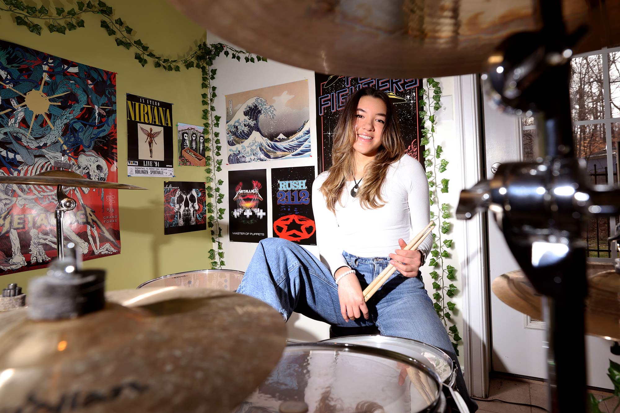 Heraldo sitting at her drumset peering between the cymbals at the photographer