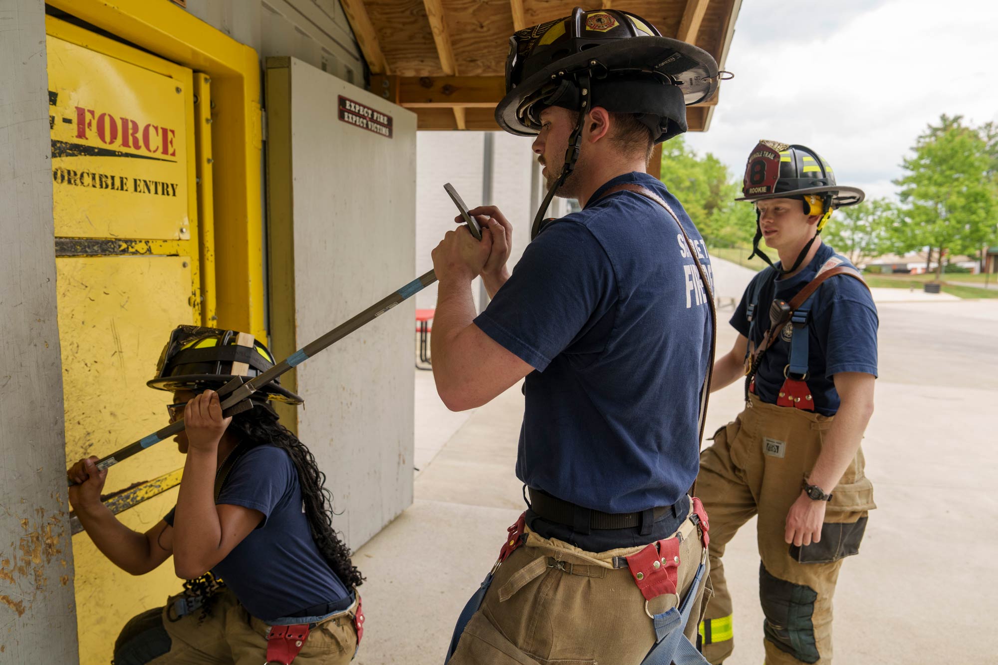 Portrait of Jones and Green training to force open doors, while Holcombe takes notes during their firefighter volunteer training.
