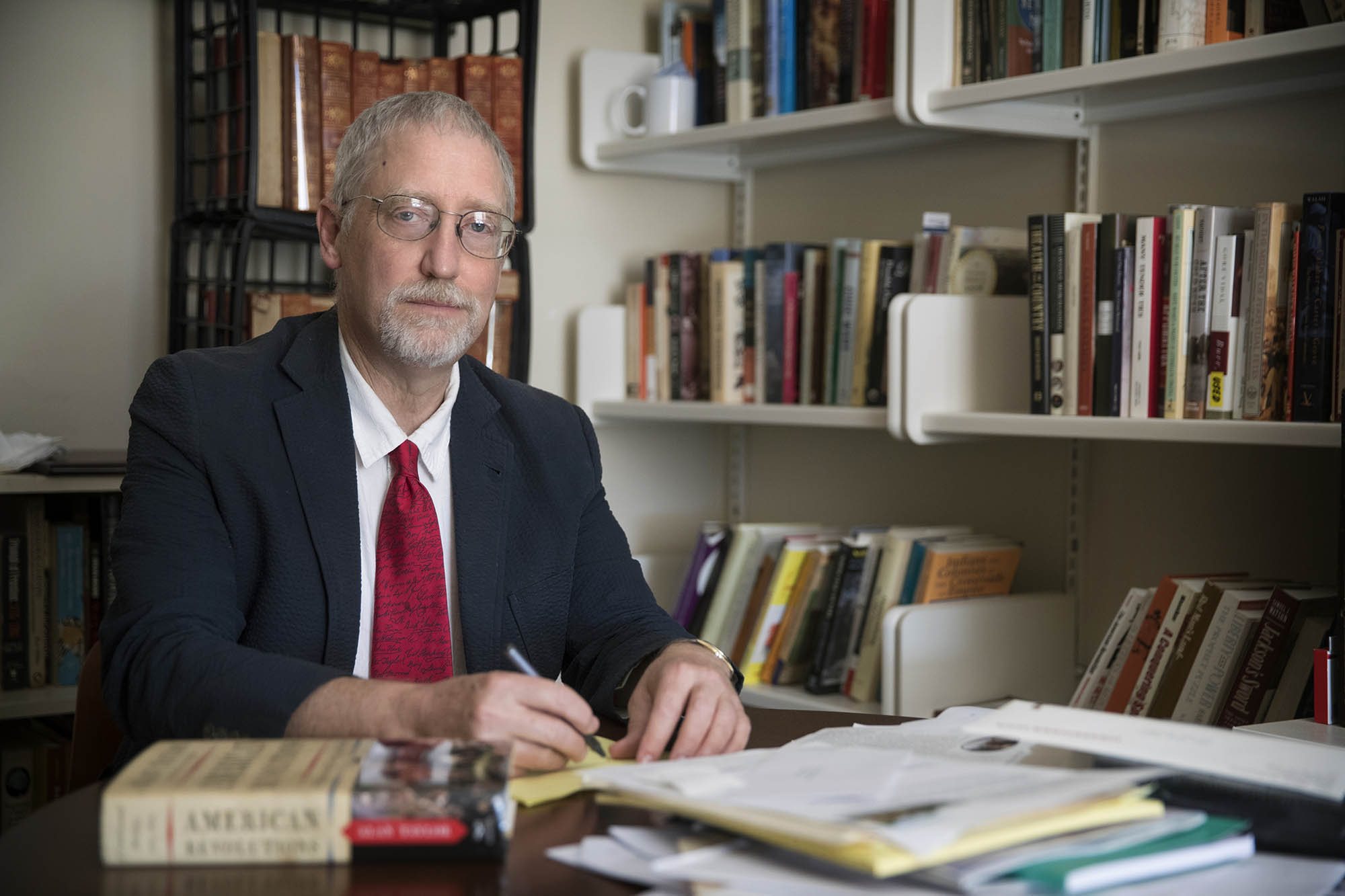 Alan Taylor sitting in a library writing. Beside him is a copy of his book “American Revolutions”.