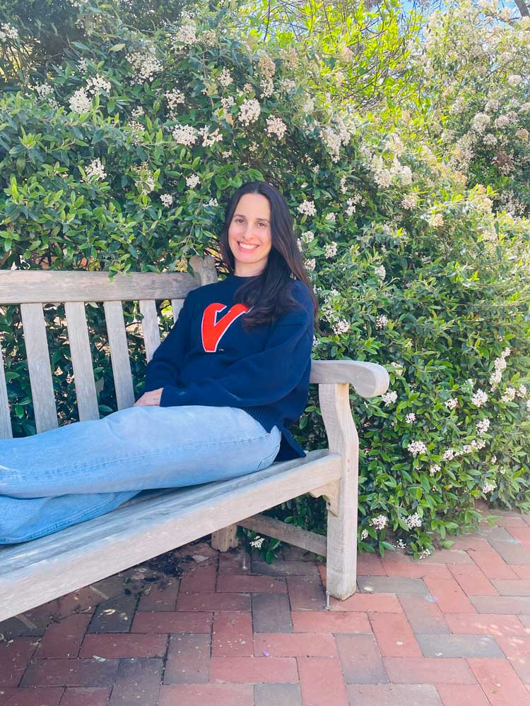 Portrait of Kathiane Schinelli sitting on a chair, stretching her leg, with a flower tree in the background.