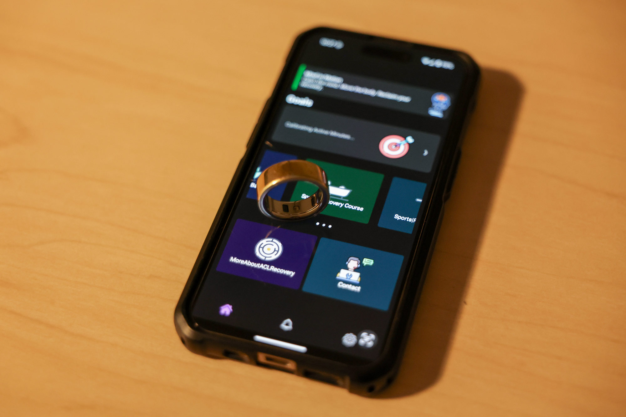 a smartphone on a desk with a digital wellness app on the screen. On the phone is placed a ring device that measures the wearer's vitals. 