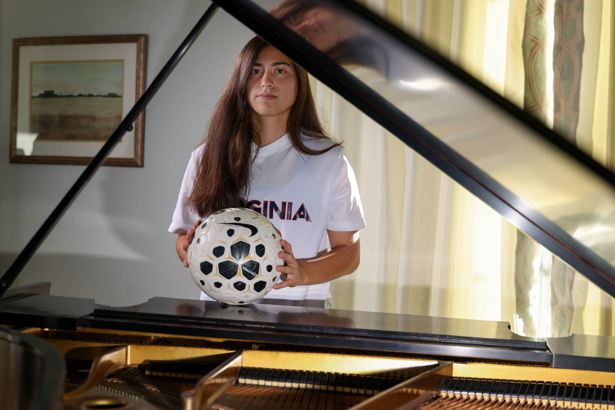 Godfrey standing behind a grand piano in her UVA soccer uniform holding a soccer ball on the lid of the piano.