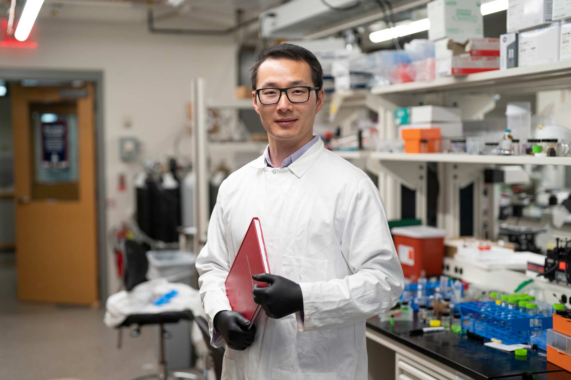 Liheng Cai standing in a laboratory holding a red notebook