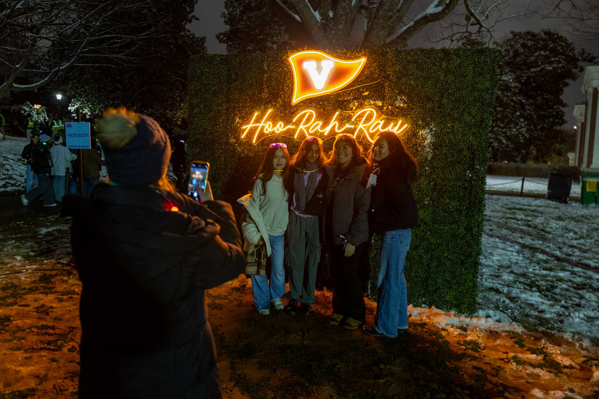 students posing in front of a photo booth backdrop with a neon sign of a 'V' flag and text reading Hoo-Rah-Ray.