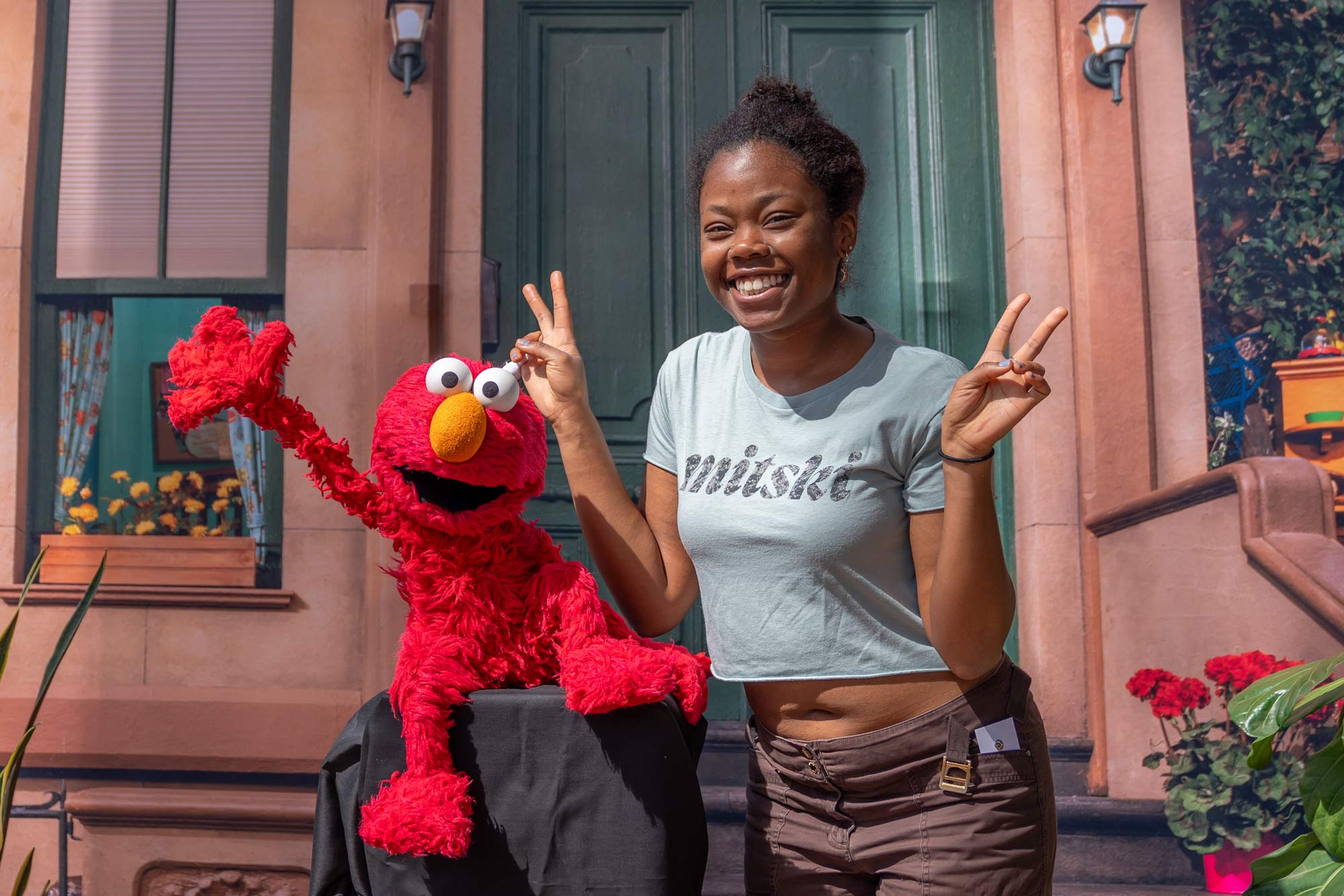 A smiling woman poses with Elmo, both flashing peace signs in front of a Sesame Street backdrop.