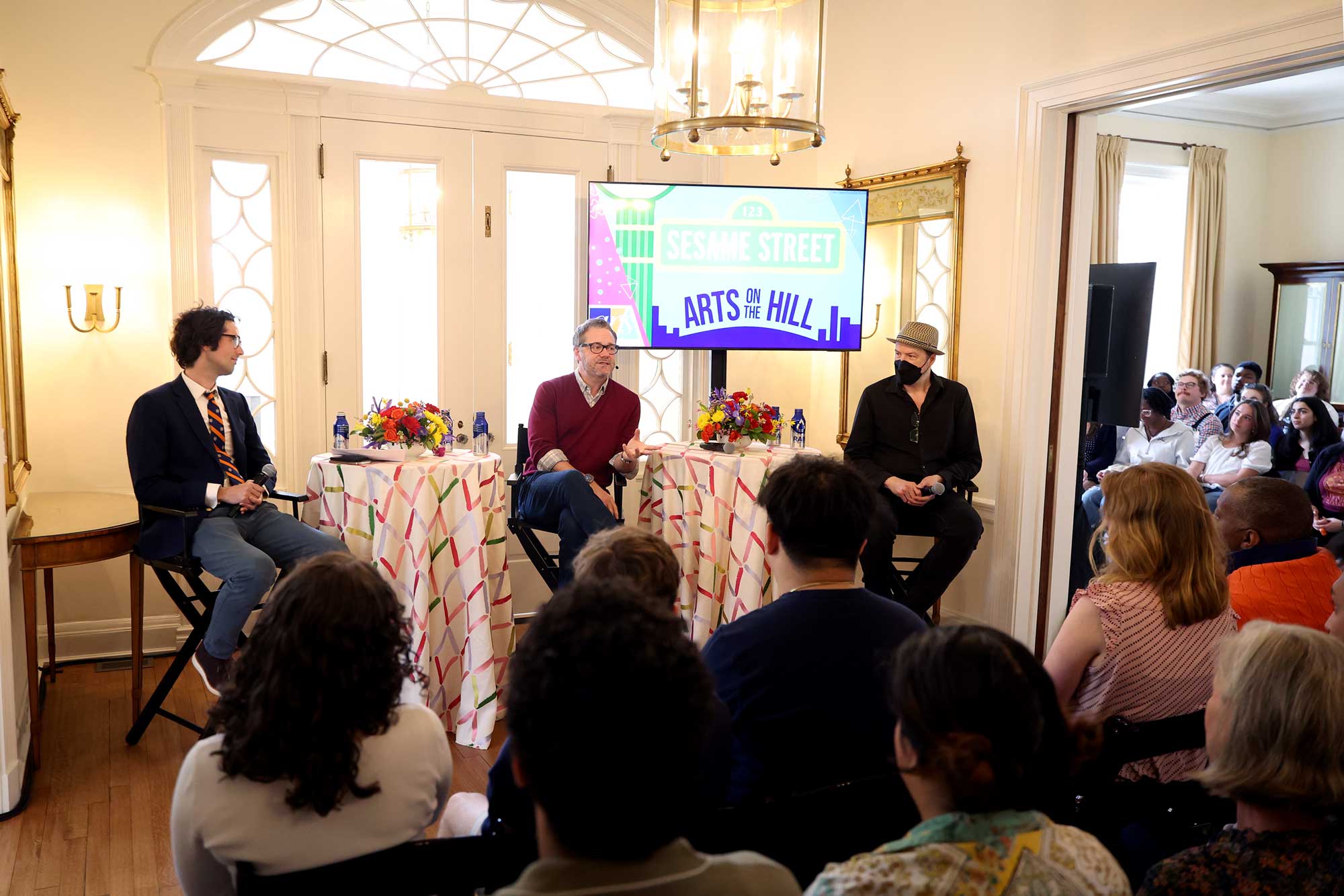 Three panelists discuss Sesame Street at an 'Arts on the Hill' event, with an engaged audience listening.