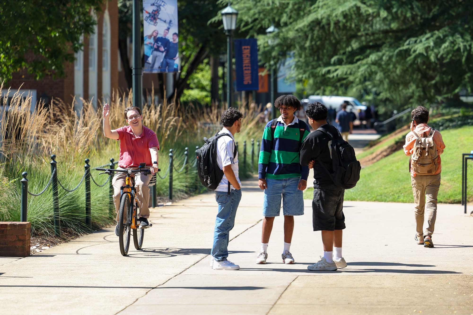 A student on a bicycle waves at the camera. Three students talk between classes. One student walks and talks on their mobile device.