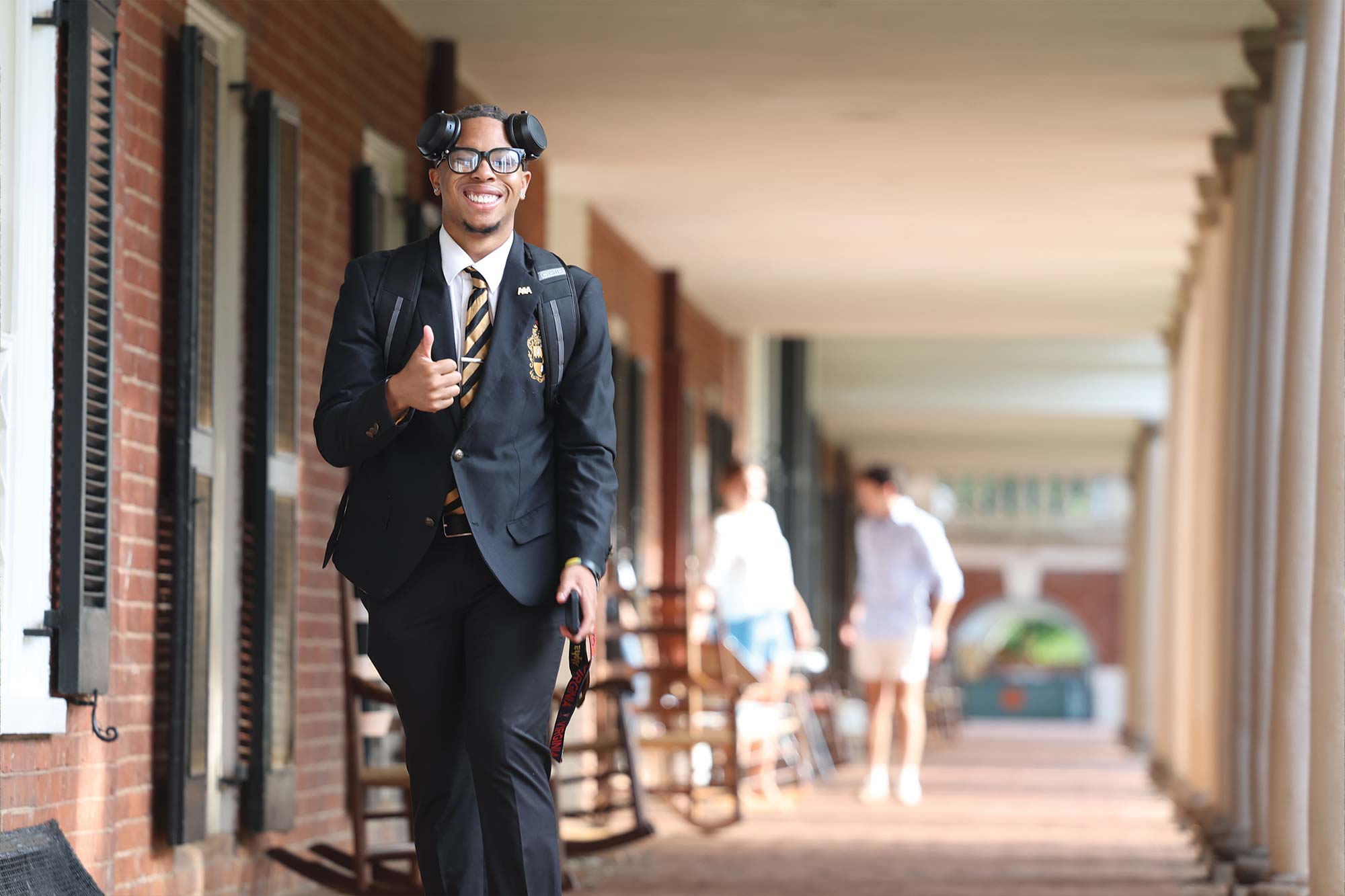 Student in a suit and headphones smiles and gives a thumbs-up in a campus hallway.