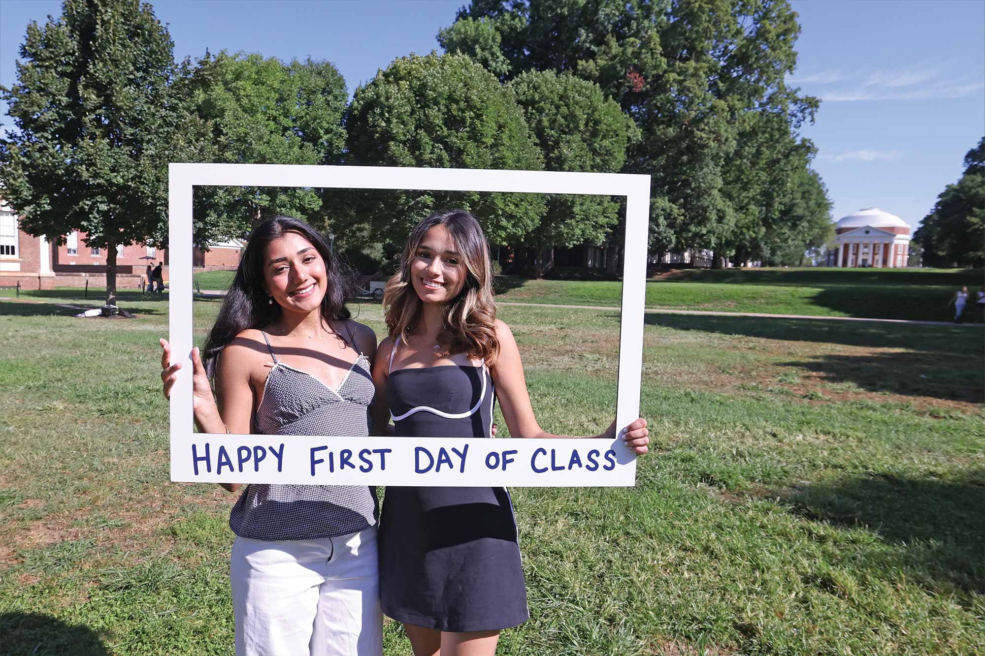 Two women smiling and holding a “Happy First Day of Class” sign on campus.