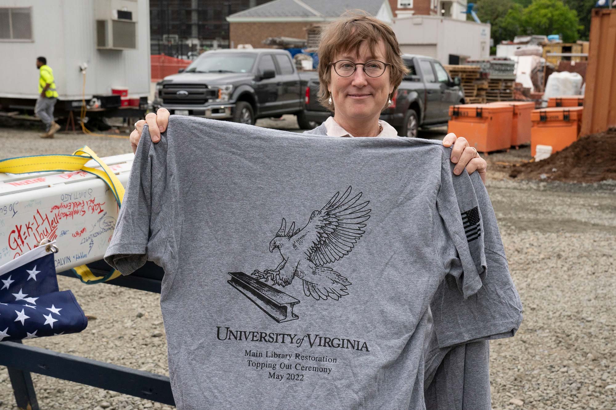 On a construction site, a woman holds up a t-shirt with an illustration of a hawk holding a steel beam