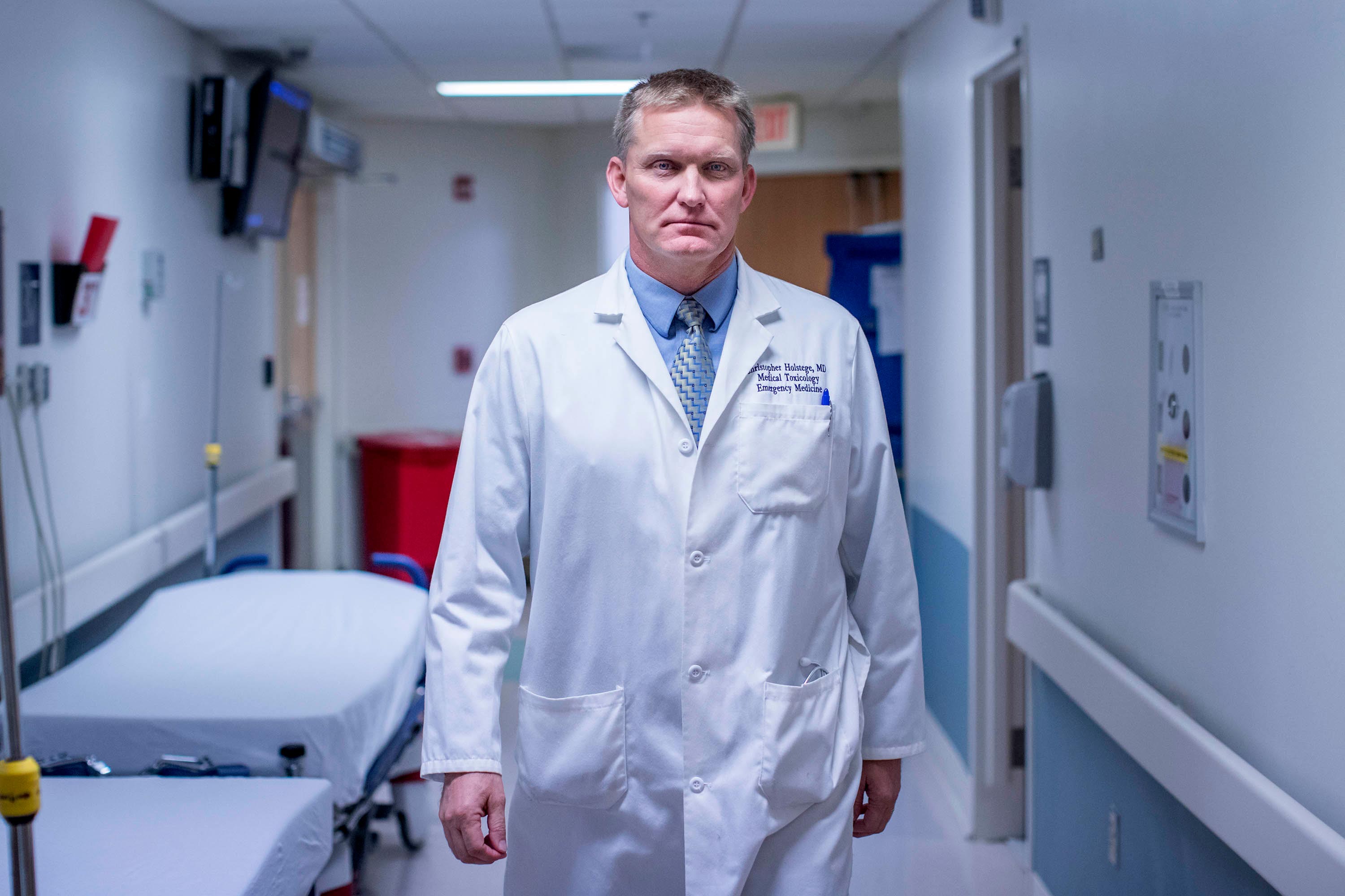 Chris Holstege in a white coat, poses for a portrait in an emergency room hallway