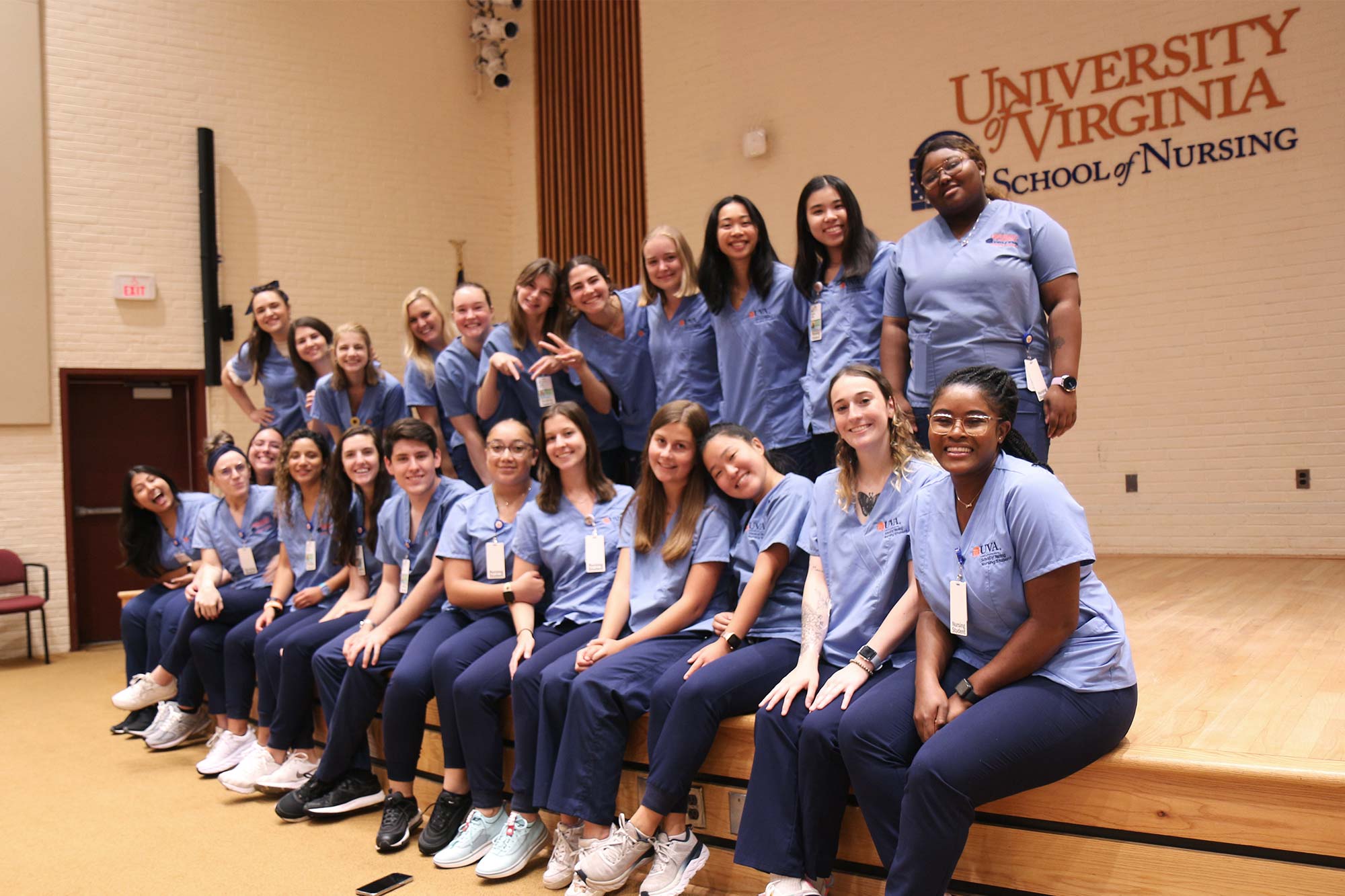 Nursing students sit and stand on a low-rise stage wearing their scrubs in front of a wall that reads "University of Virginia, School of Nursing"