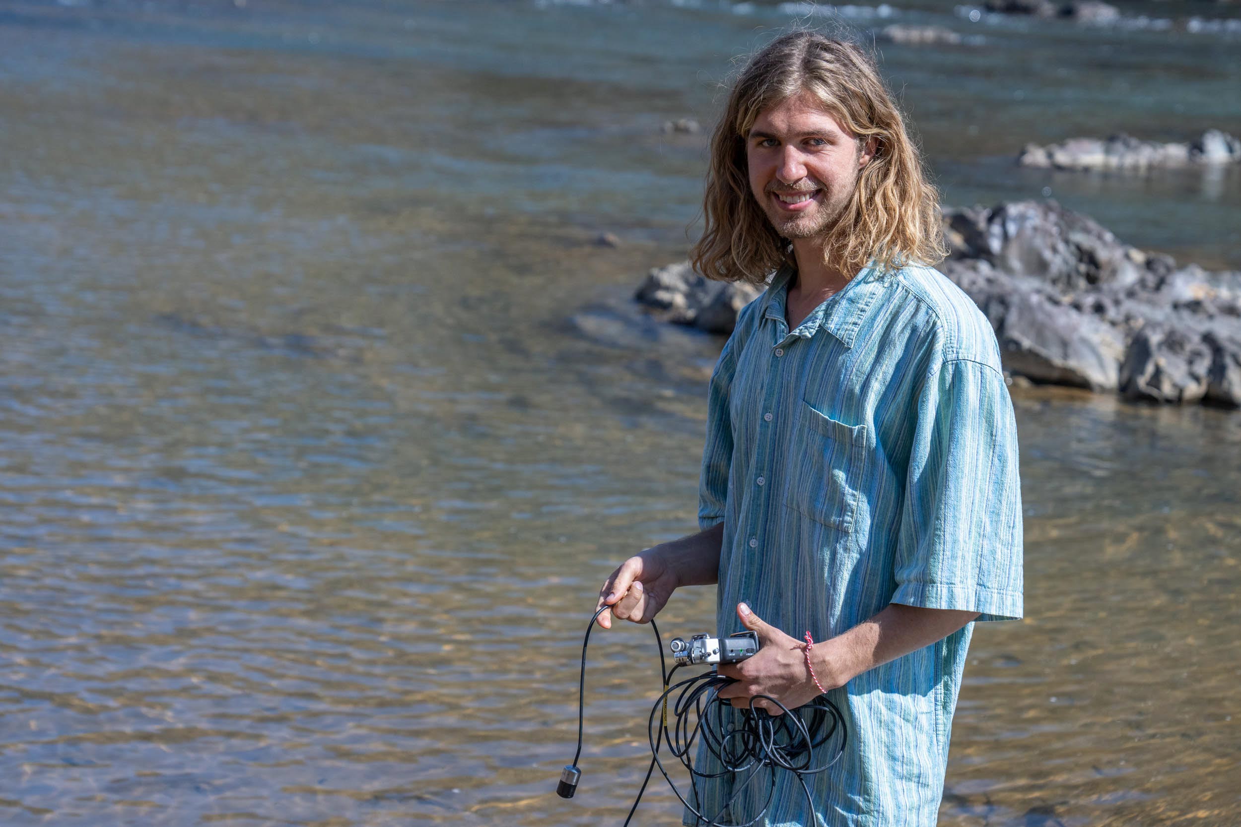 Davis Coffey stands in a stream, holding a device and a mass of cables, and smiles at the camera