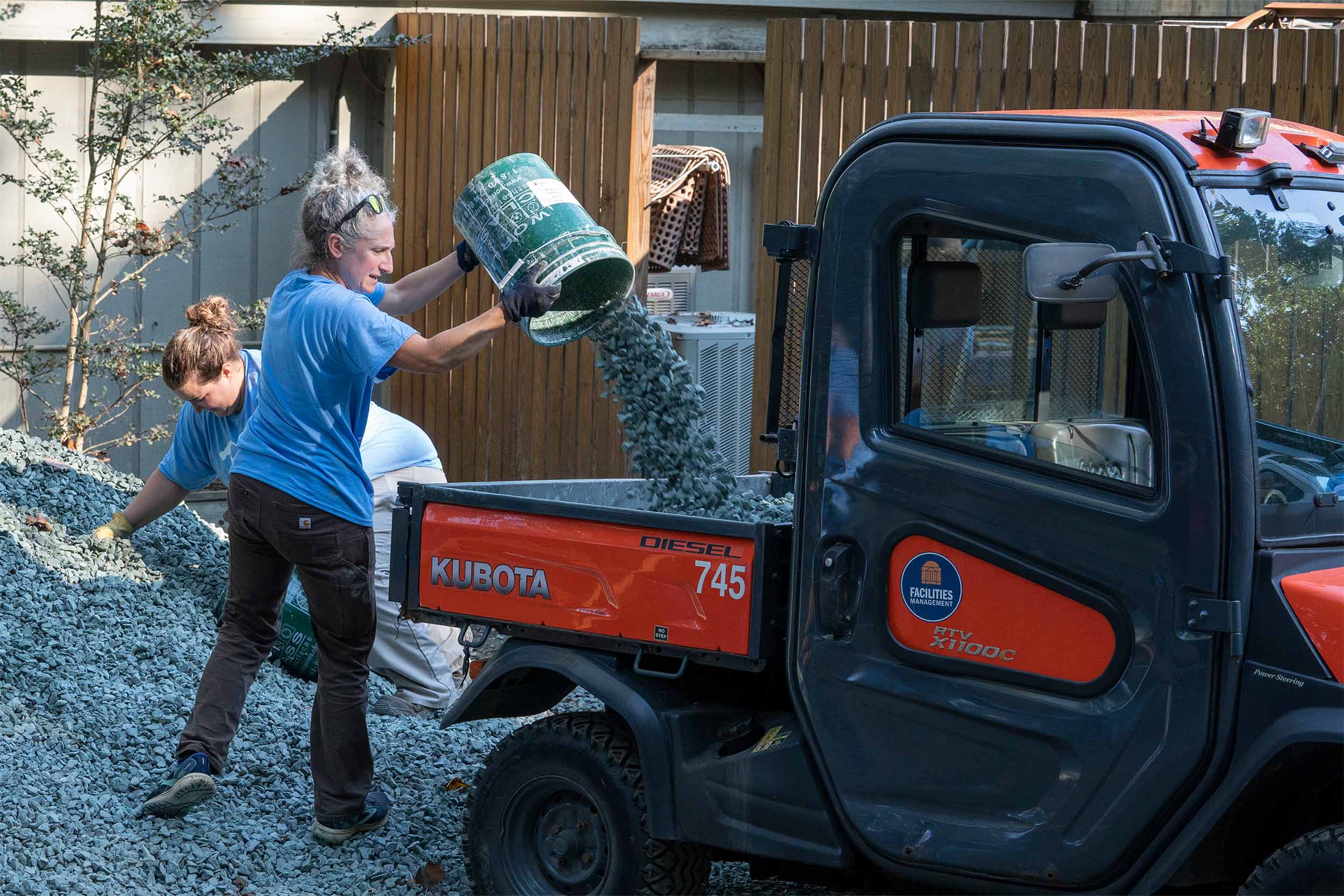 A woman dumps a bucket of gravel into the bed of a utility vehicle.
