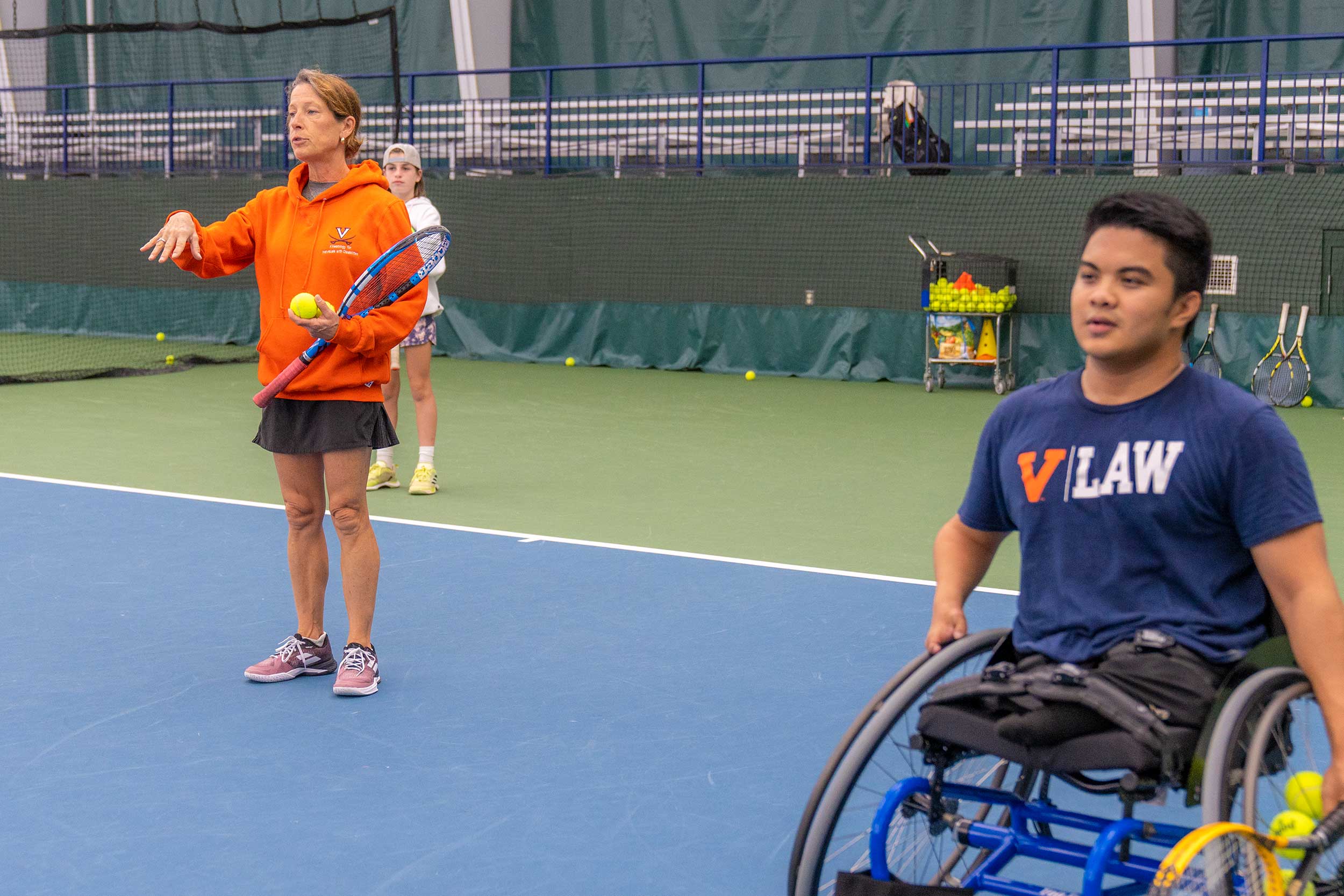 Woman in orange UVA sweatshirt, while holding a racket, provides instruction at a wheelchair tennis practice. In foreground is UVA student in a wheelchair.