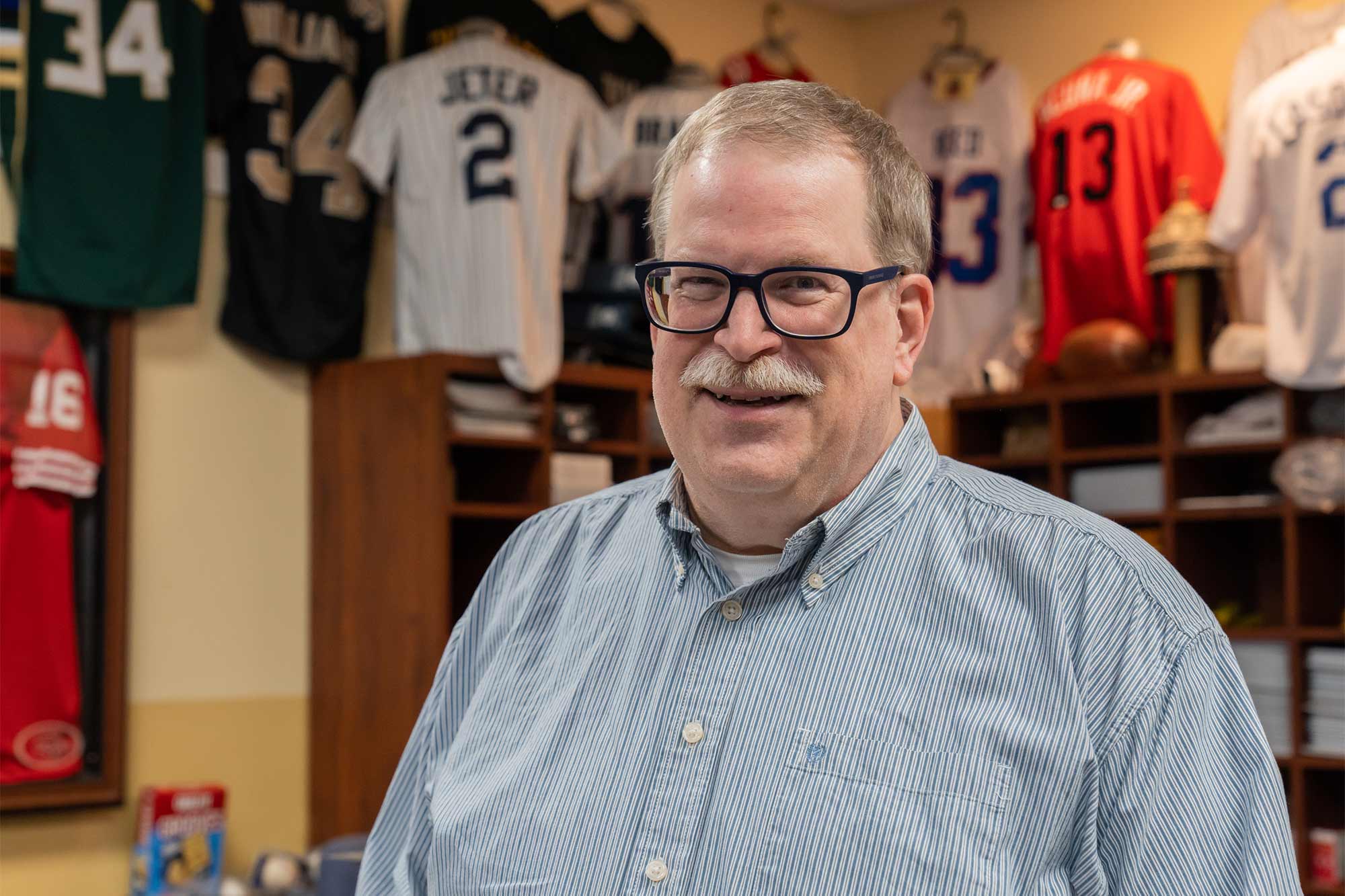 Jeff Prillaman stands in his shop in front of shelving units and multiple sports jerseys hanging on the wall