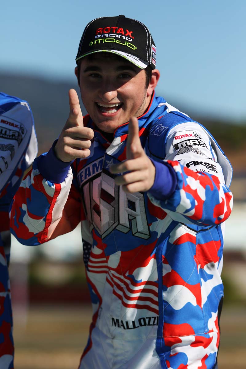 A man in racing gear smiles and points at the camera with his thumbs in the air