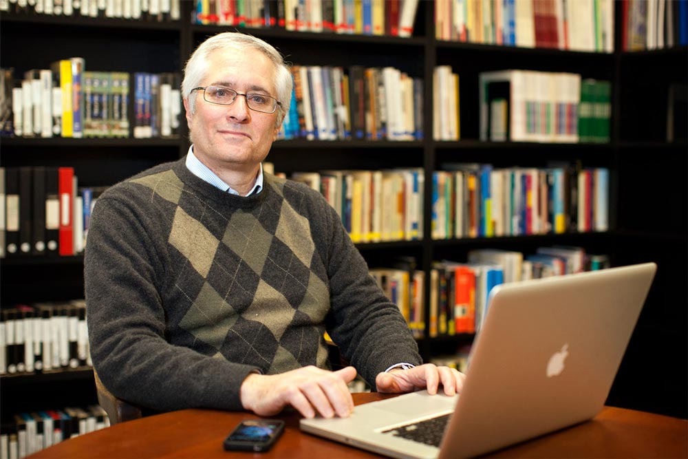 Bruce Williams sits at a laptop in a library and smiles at the camera