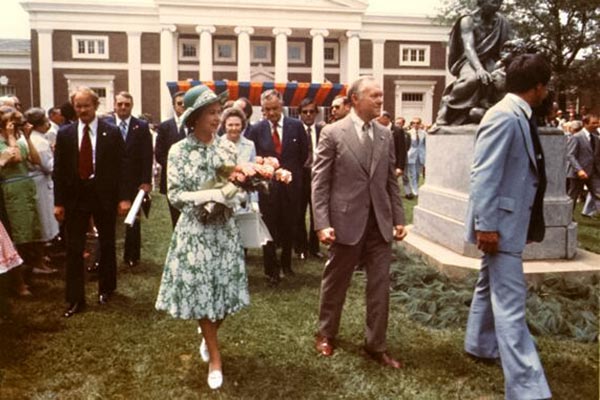 Queen Elizabeth holds a bouquet of roses and walks down the lawn with a group of stylishly dressed people