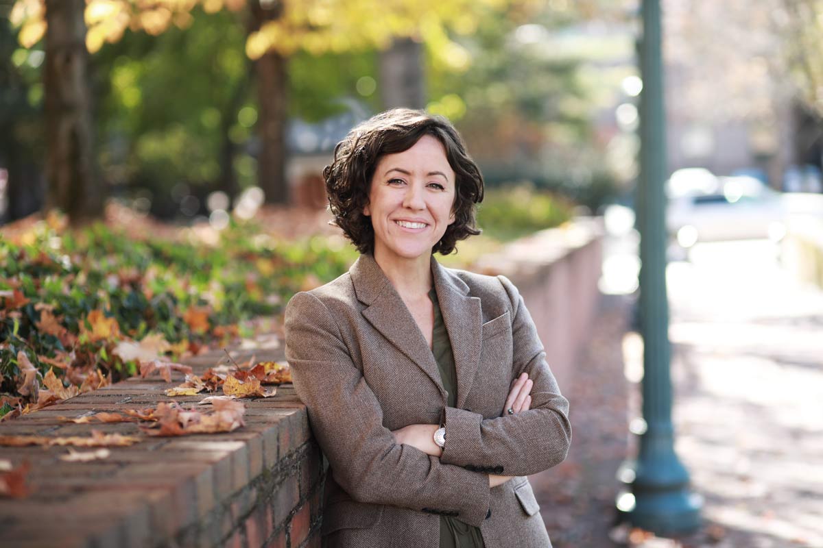 Professor Kelly leans against a brick wall with her arms crossed as she smiles at the camera. 