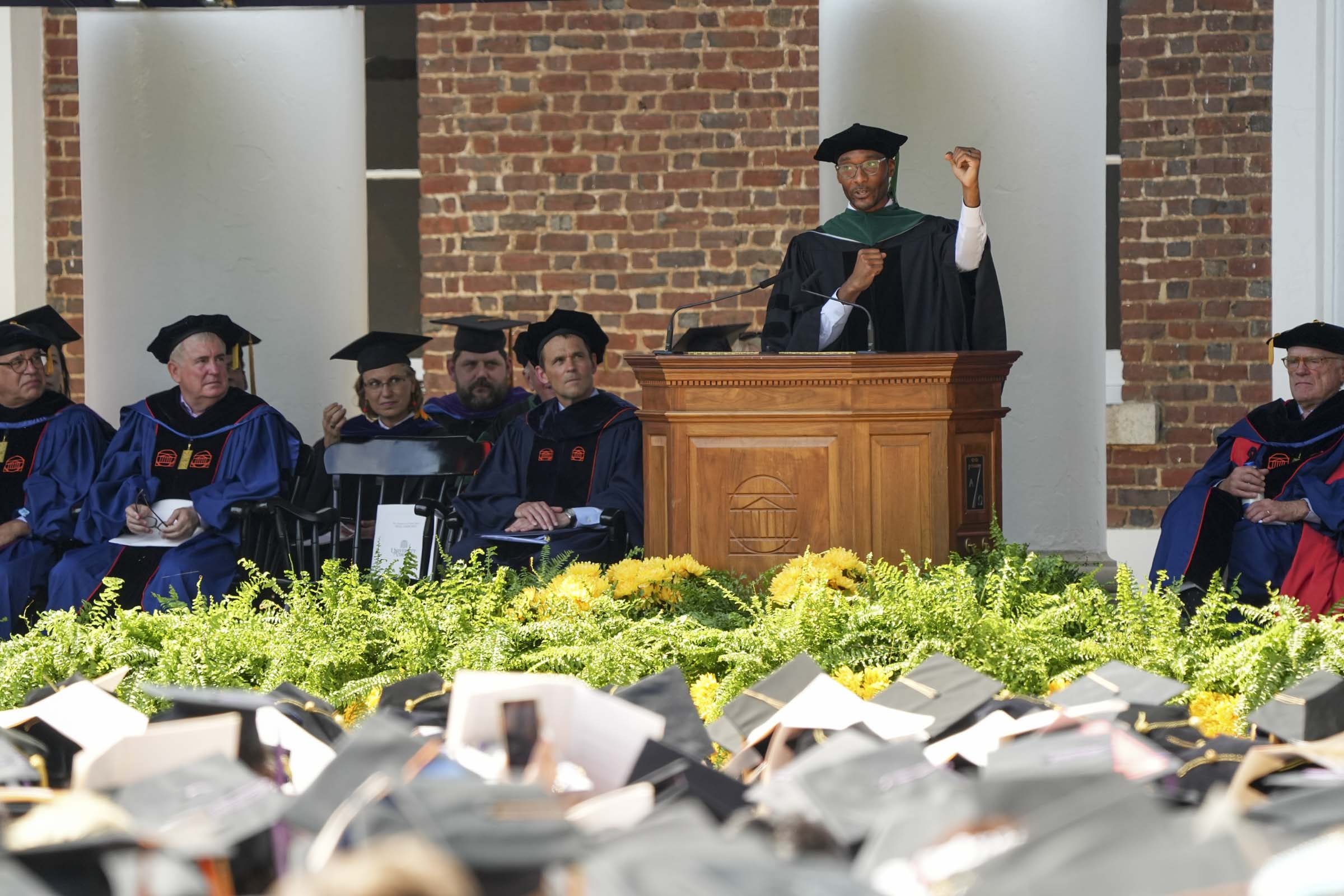 Taison Bell, speaking from a podium to a crowd of seated graduates in caps, gestures with closed fists