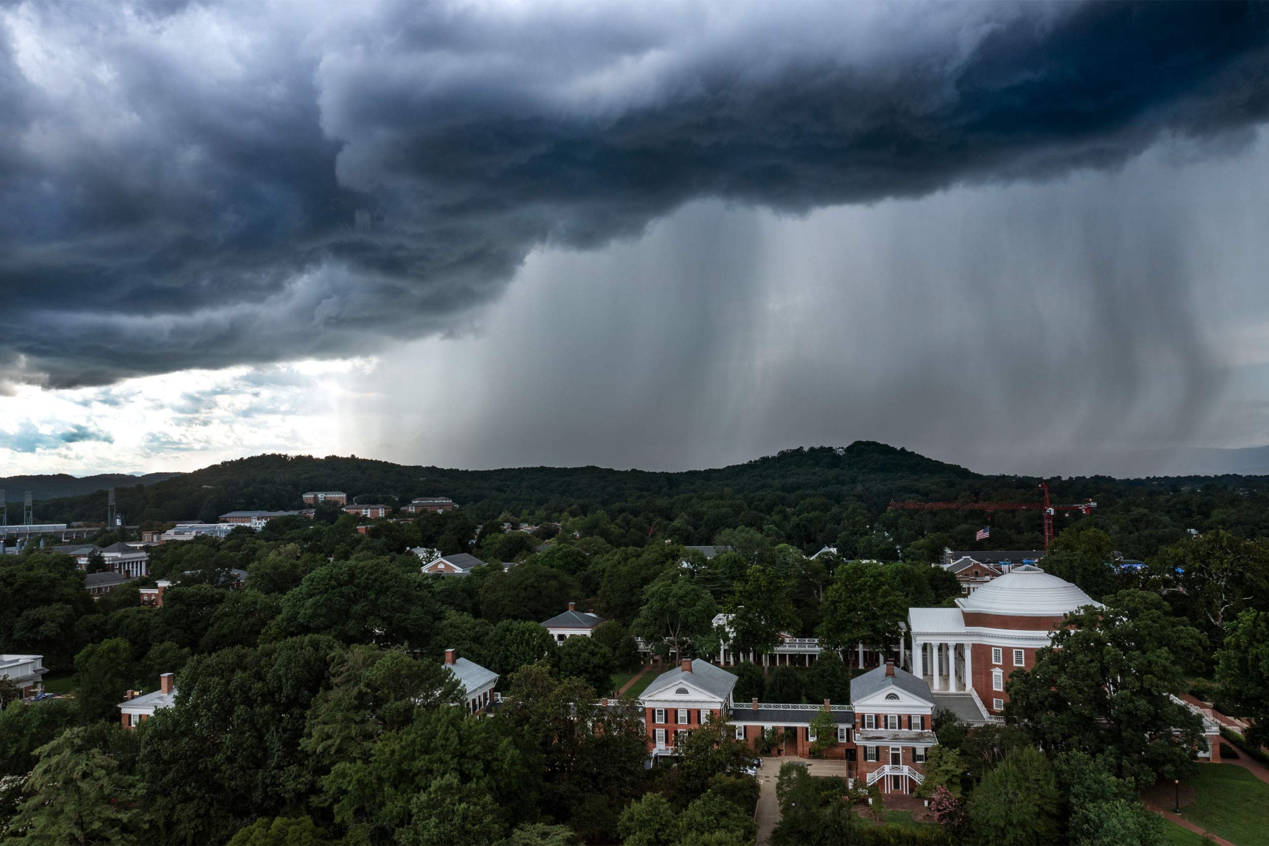 An aerial view of UVA Grounds, featuring the Rotunda on the right side, with dark clouds covering the sky. A wall of rain looms in the distance. 