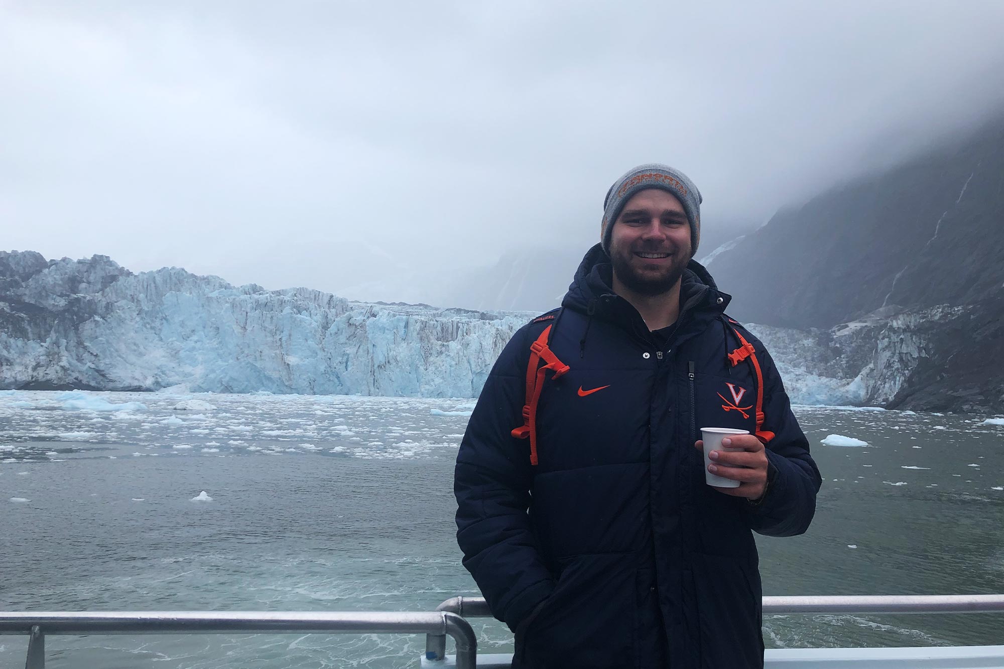 Pleško stands in front of glaciers holding a cup and wearing a UVA winter coat and a greay beanie